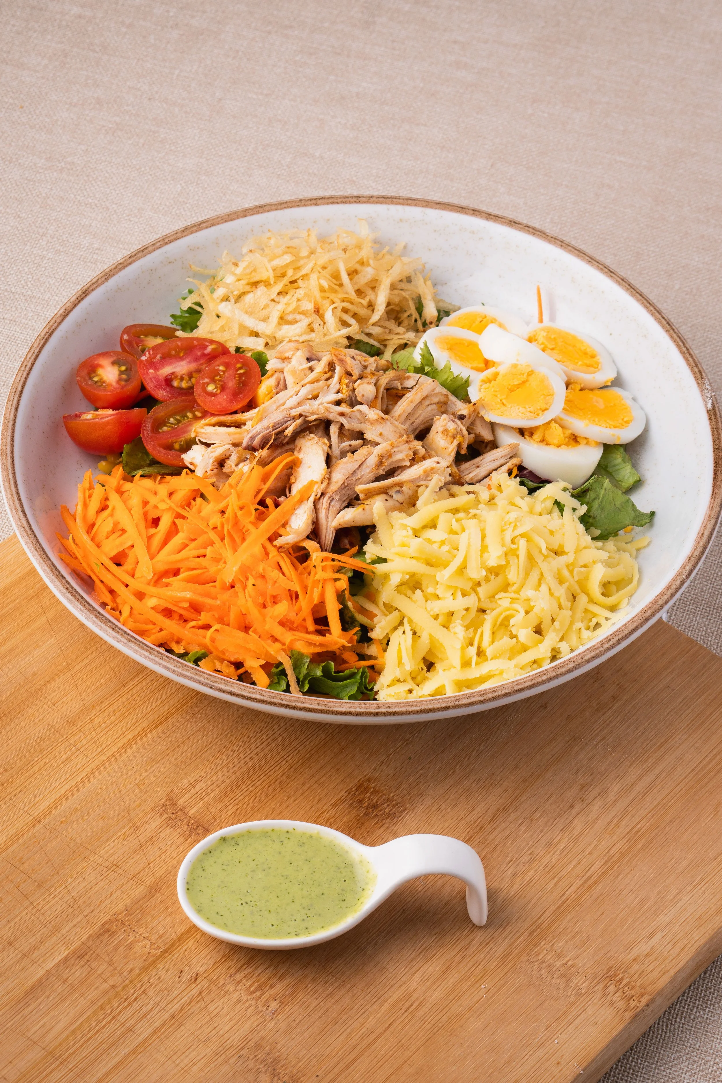 A salad bowl with cherry tomatoes, shredded carrots, shredded cheese, shredded chicken, sliced hard-boiled eggs, shredded chicken, and shredded cheese, served with a side of green dressing on a small white dish, placed on a wooden board.