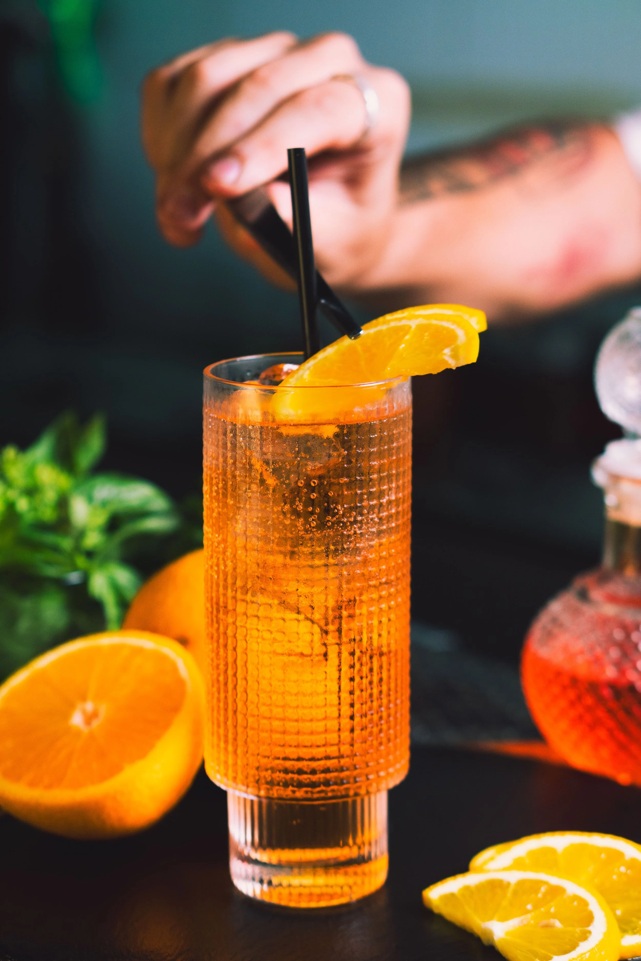 A tall glass of orange-colored drink garnished with an orange wedge and a black straw, with lemons and a person’s hand stirring in the background.
