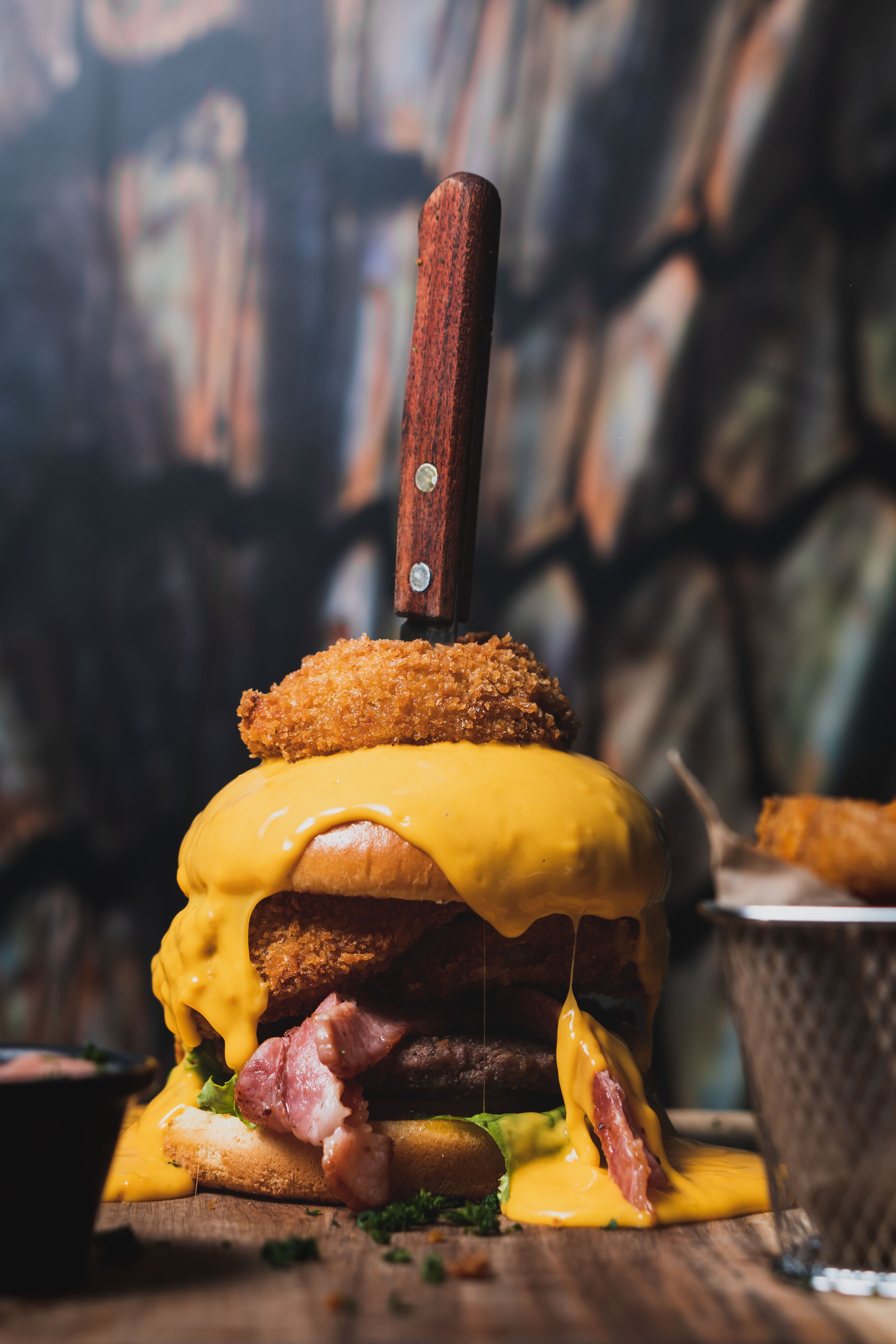 A towering cheeseburger with melted cheese, fried chicken, bacon, and a knife in a rustic setting.