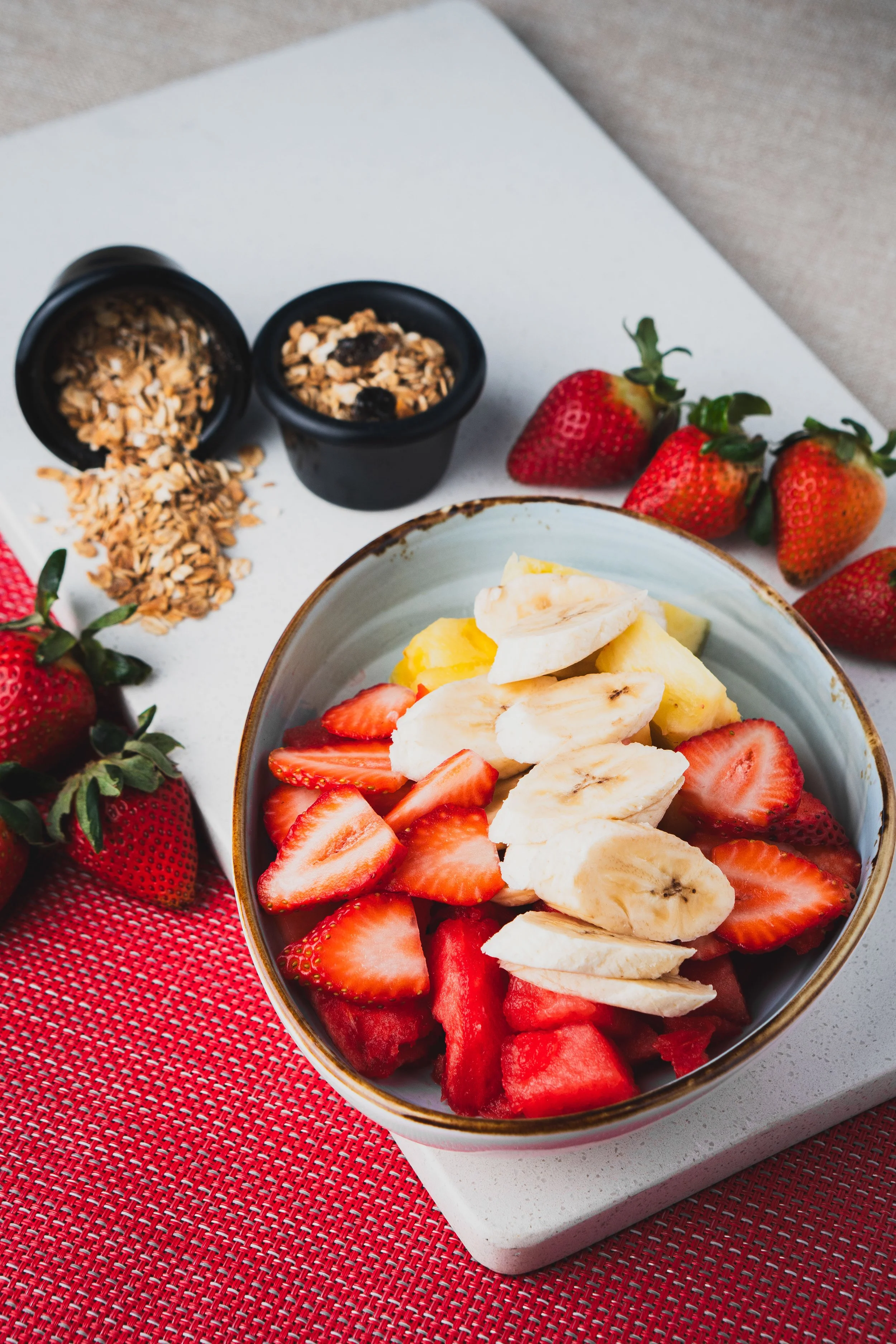 A bowl of sliced strawberries, bananas, and watermelon on a white surface with fresh strawberries and granola in small black containers nearby.