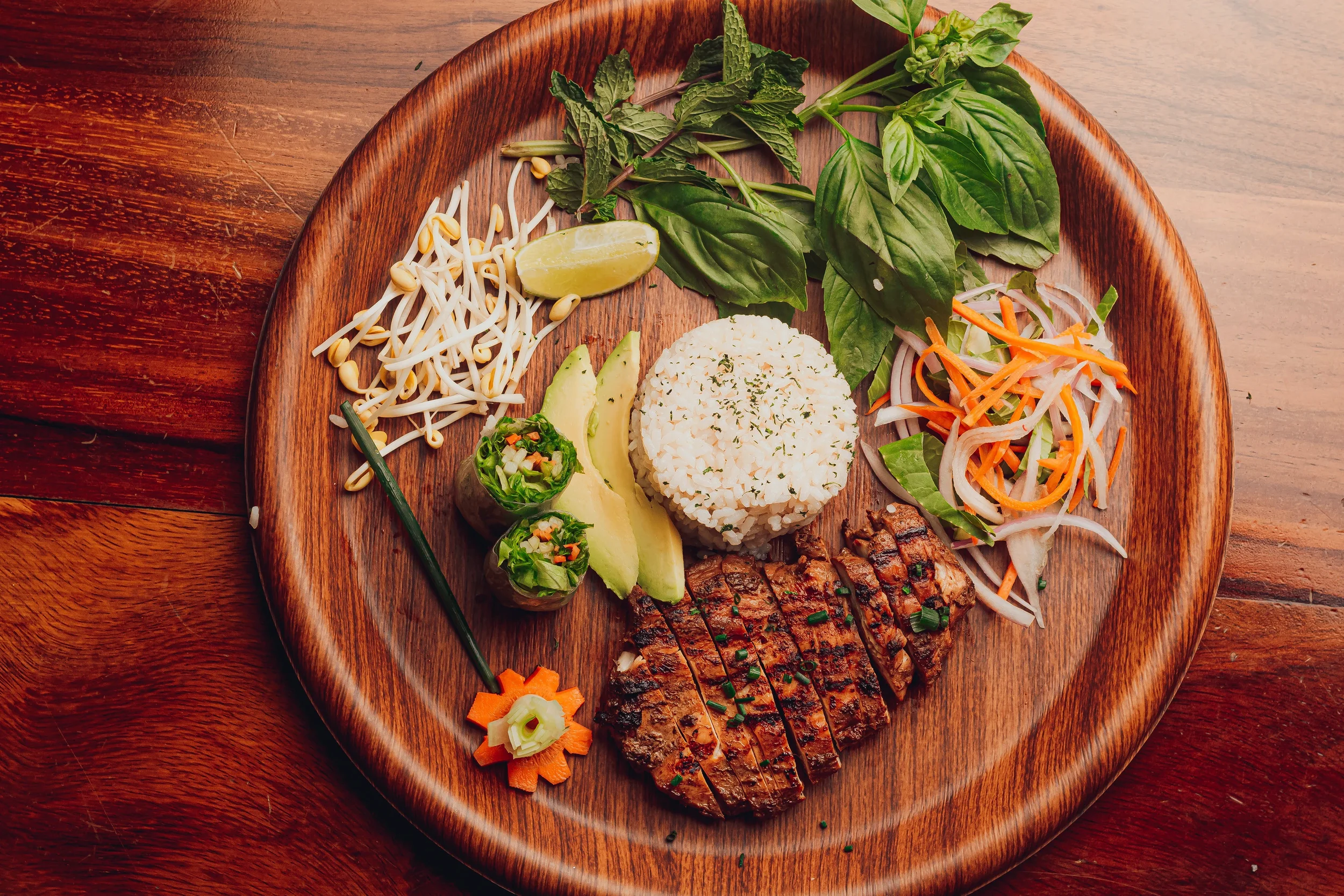 A wooden plate holds Asian-inspired food including grilled sliced beef, sushi rolls with avocado, rice, bean sprouts, shredded carrots and onions, fresh basil, mint, a lemon wedge, and a small carved carrot flower with a green onion stalk in the cent