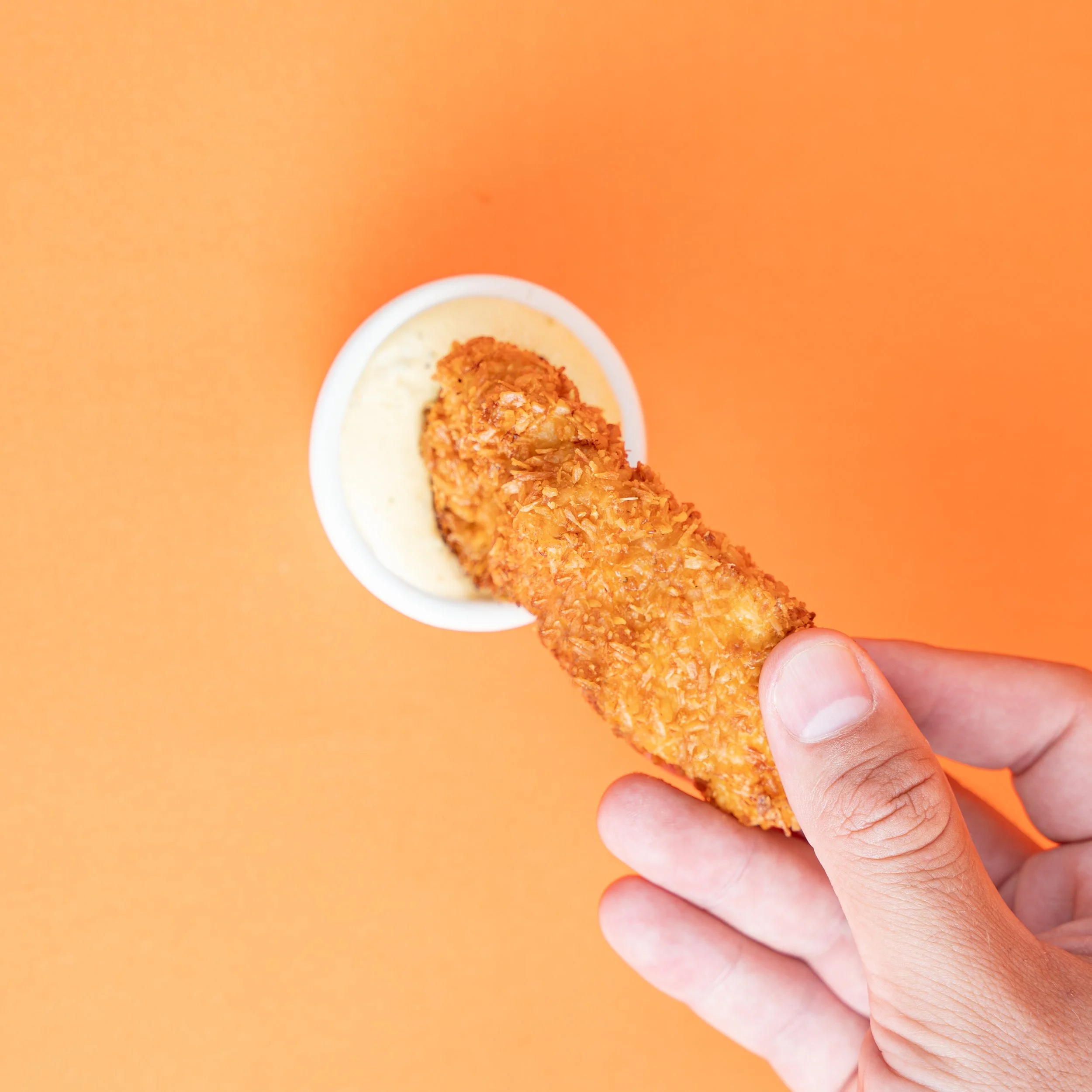 Persona sosteniendo un pescado empanizado frito con una salsa en un recipiente blanco sobre fondo naranja.