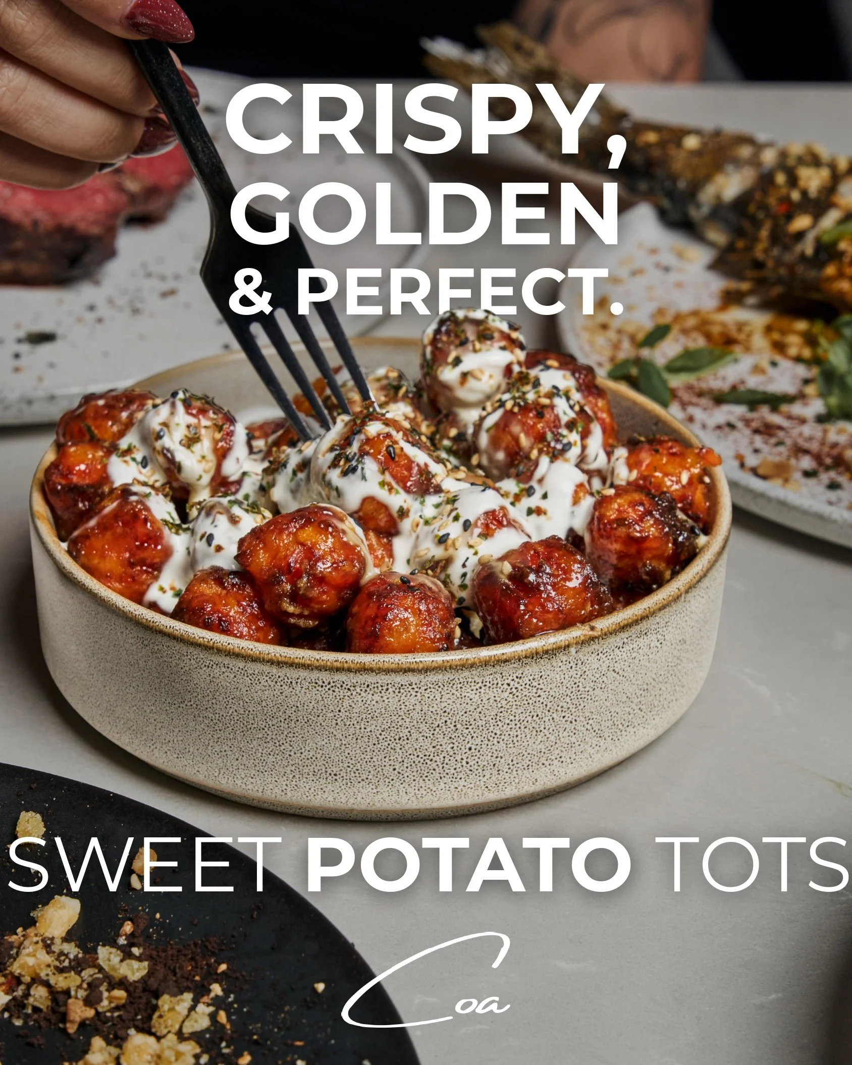 A bowl of sweet potato tots topped with sauce and herbs on a table with other dishes, with text overlay reading "Crispy, golden & perfect. Sweet potato tots".