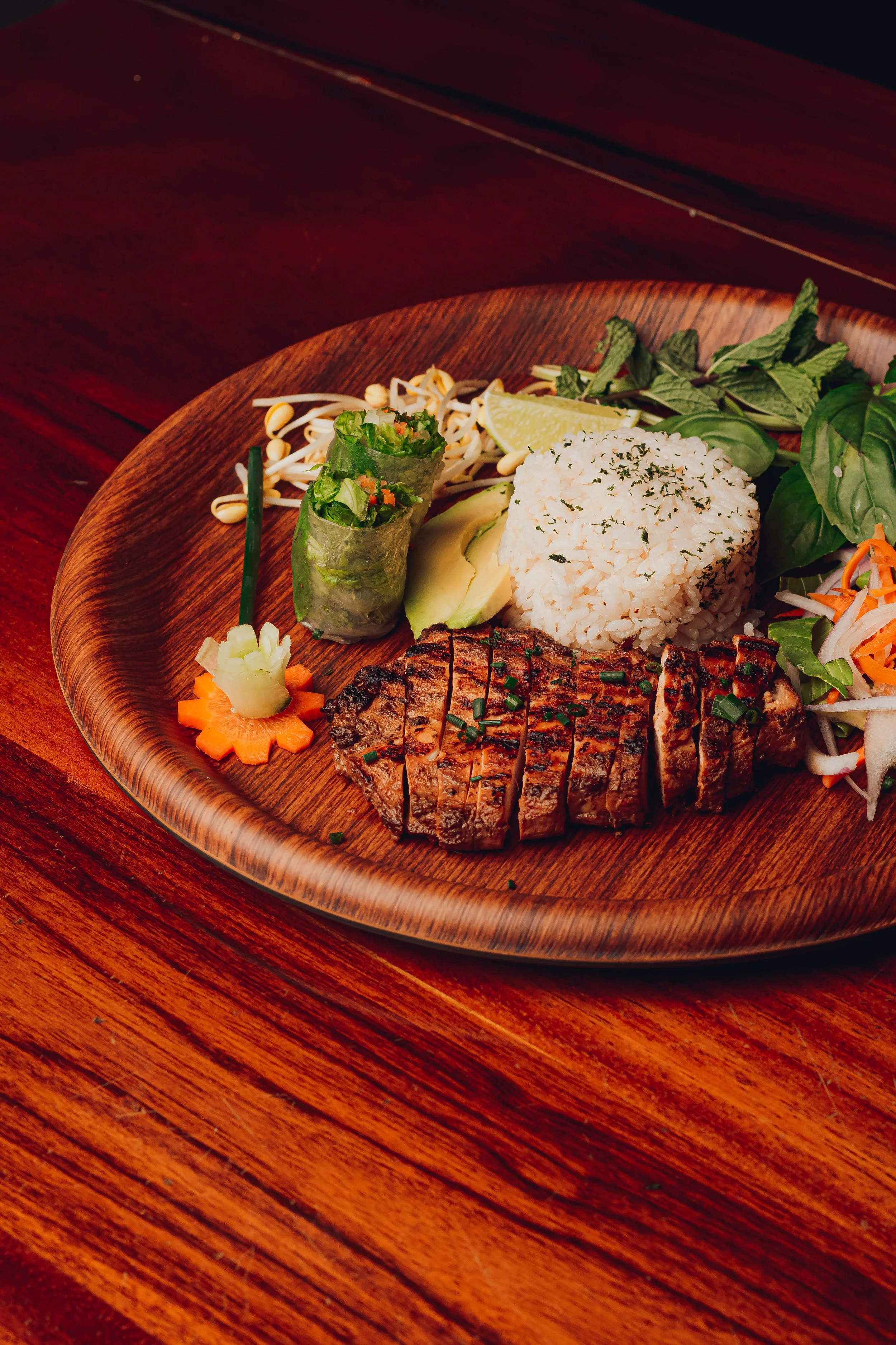 A wooden plate with grilled steak, white rice, salad, spring roll, lemon wedge, avocado, and vegetable garnishes on a wooden table.