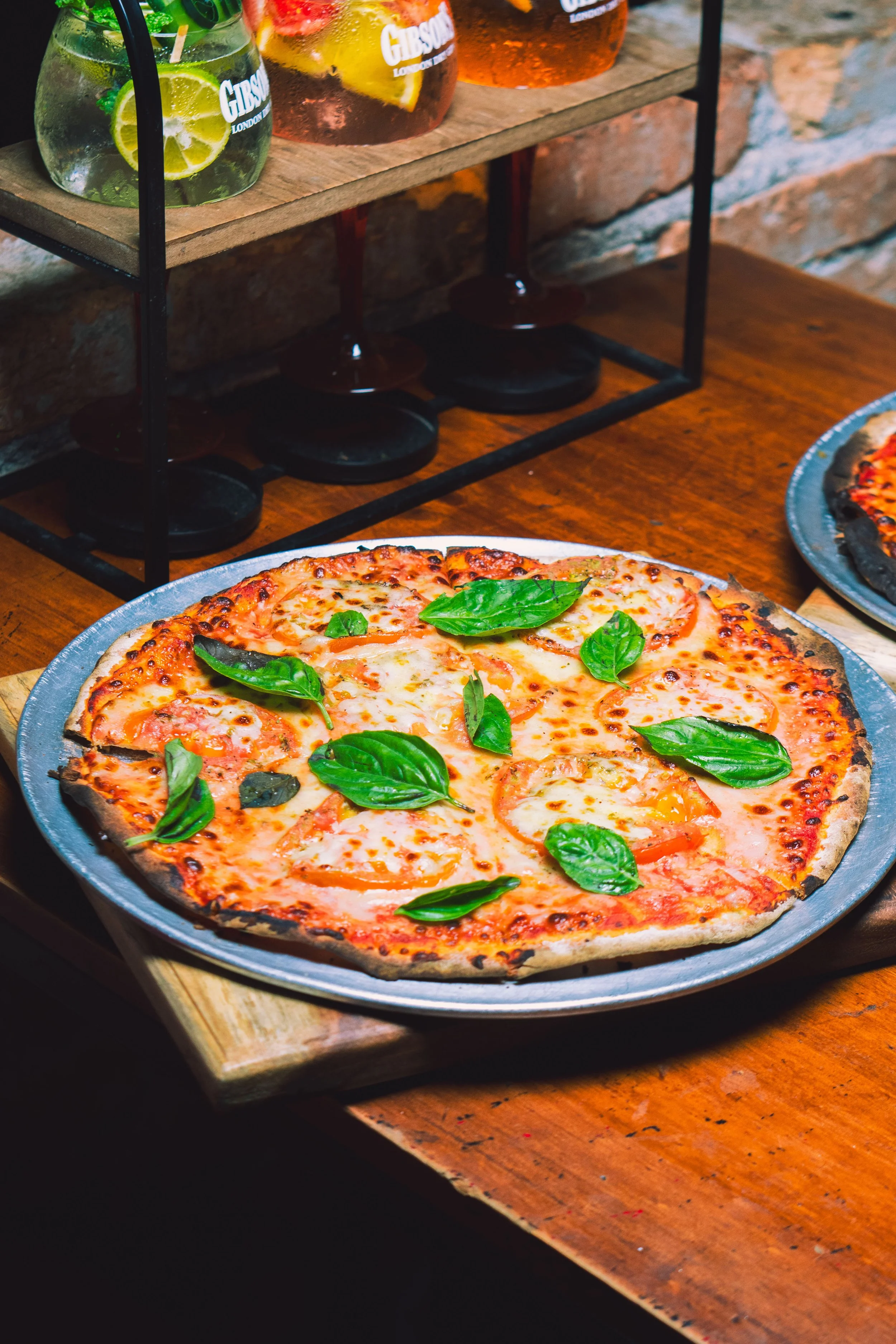 A thin-crust pizza topped with cheese, tomatoes, and fresh basil leaves, served on a metal tray on a wooden table, with a side of colorful beverage drinks behind it.