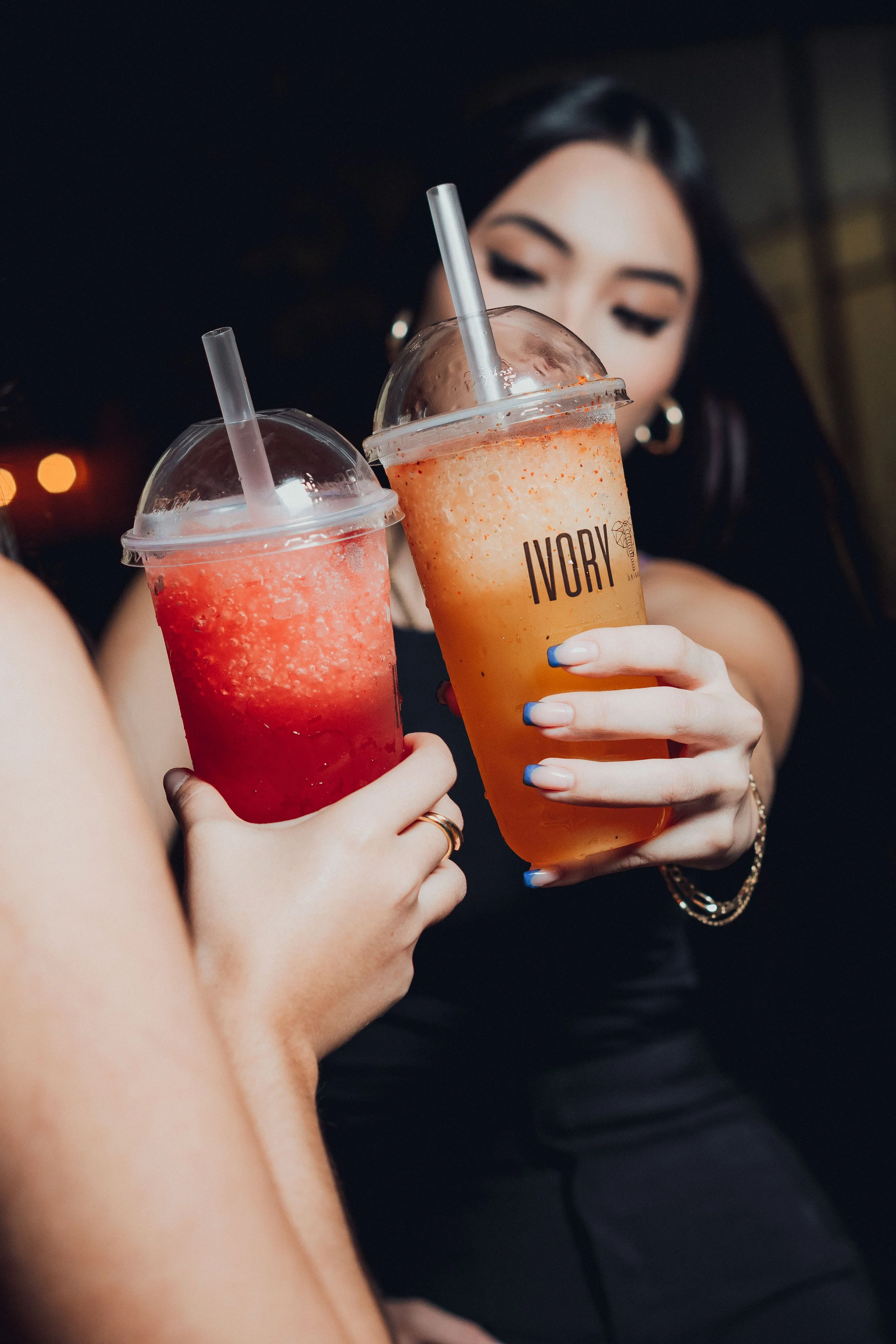 Two women holding colorful frozen drinks in plastic cups with straws, one red and one orange, in a social setting with a dark background