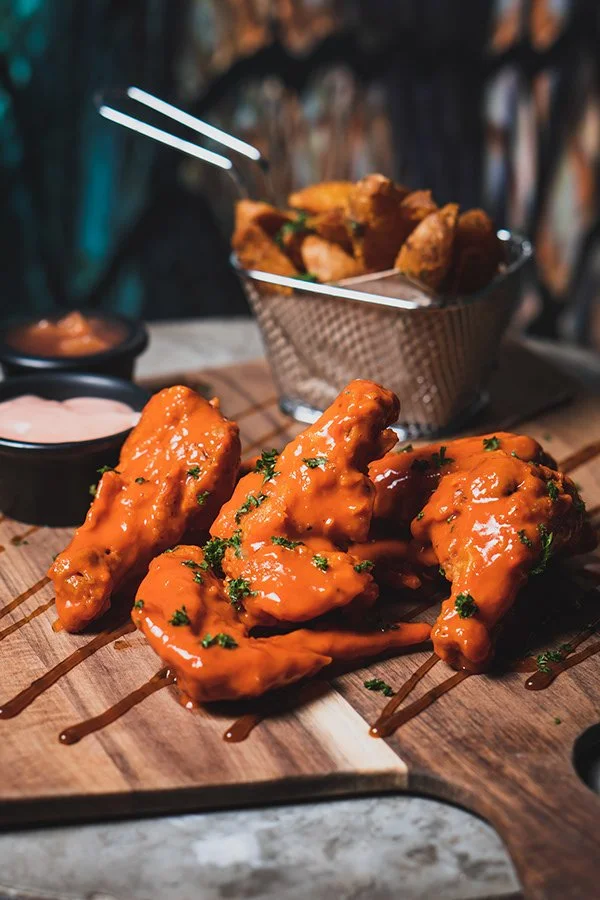 Three pieces of buffalo chicken wings glazed with spicy sauce, garnished with chopped parsley, served on a wooden board with side sauces and a basket of fried chicken in the background.
