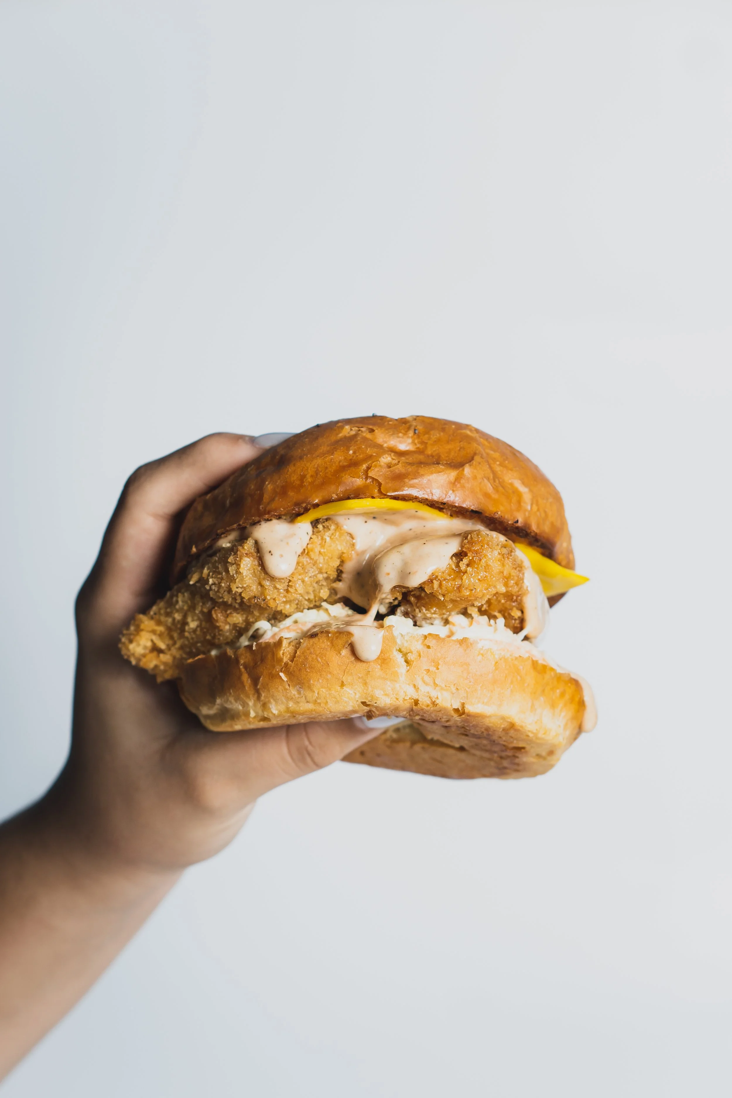 Hand holding a fried chicken sandwich with sauce, pickles, and a bun against a plain background.
