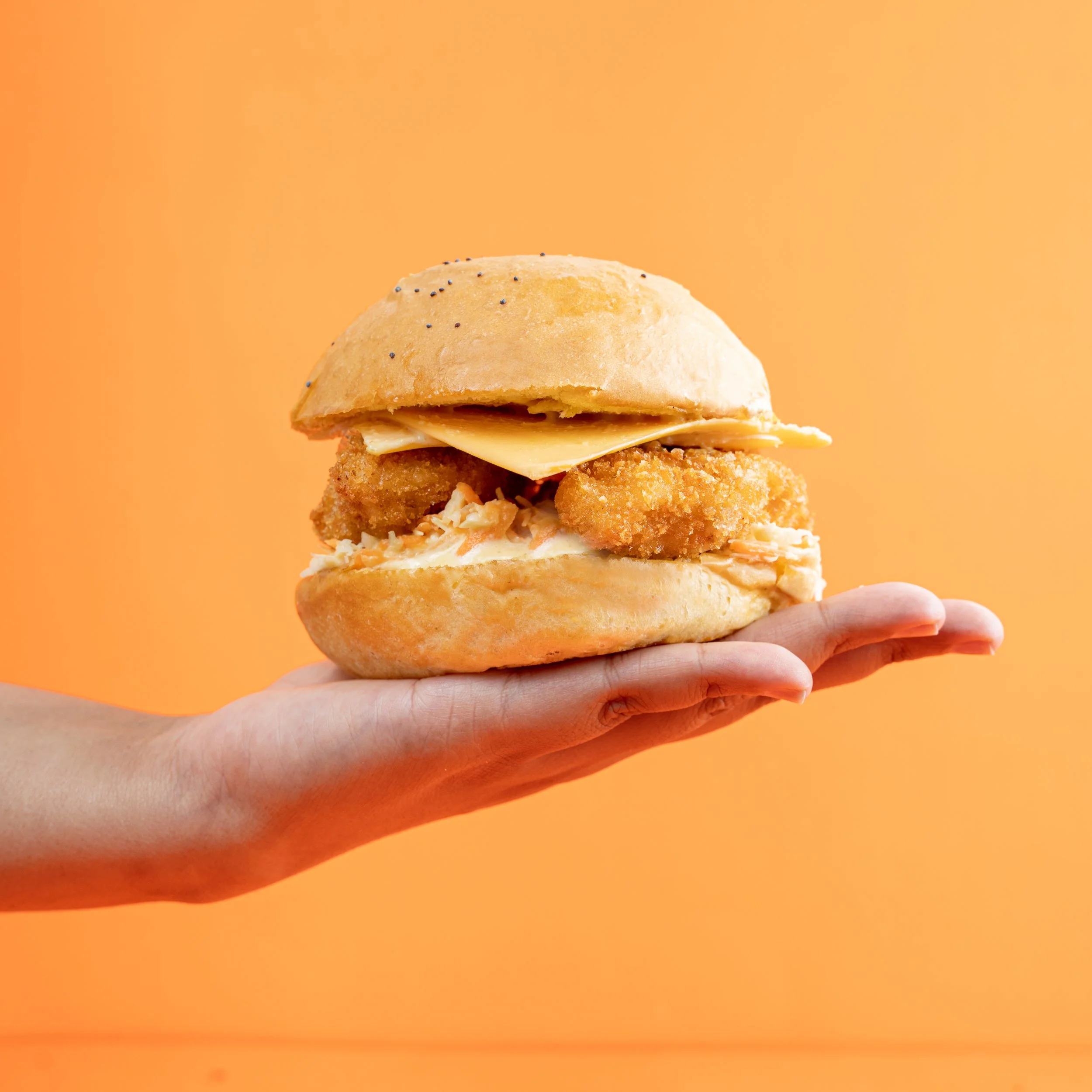A hand holding a fried fish sandwich with tartar sauce, cheese, and a sandwich bun, against an orange background.