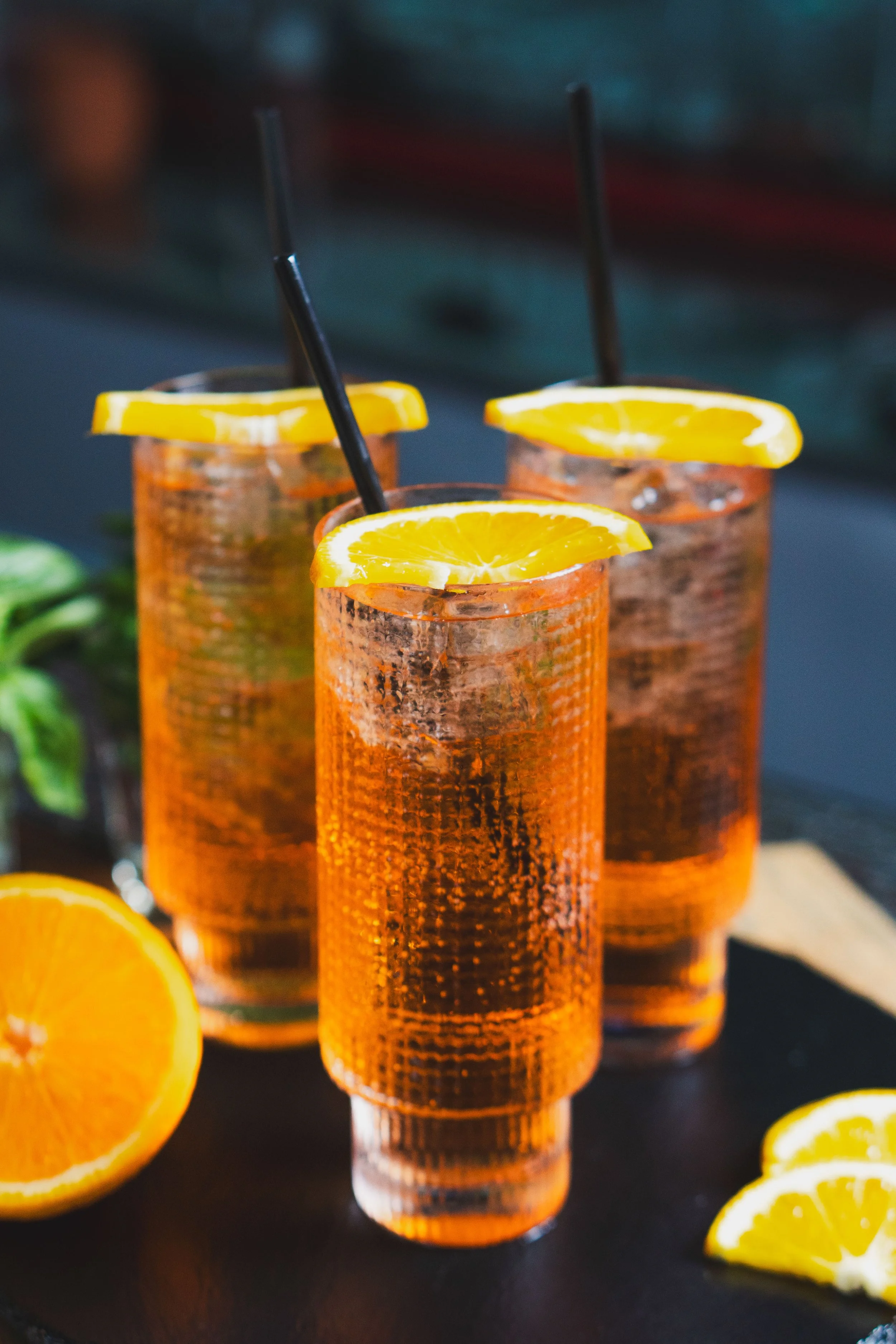 Three tall glasses filled with an orange-colored beverage, each garnished with a lemon wedge and a black straw, on a dark table with lemon slices nearby.