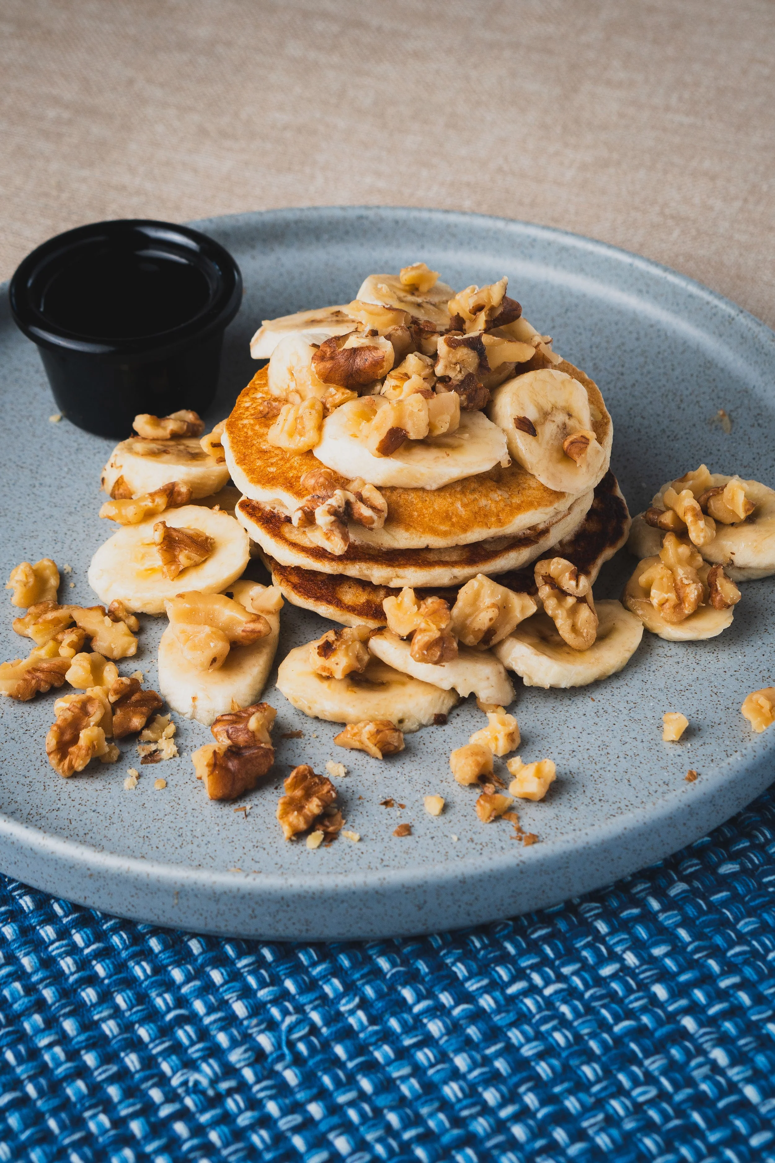 A plate of three banana pancakes topped with chopped nuts, served with a side of syrup.