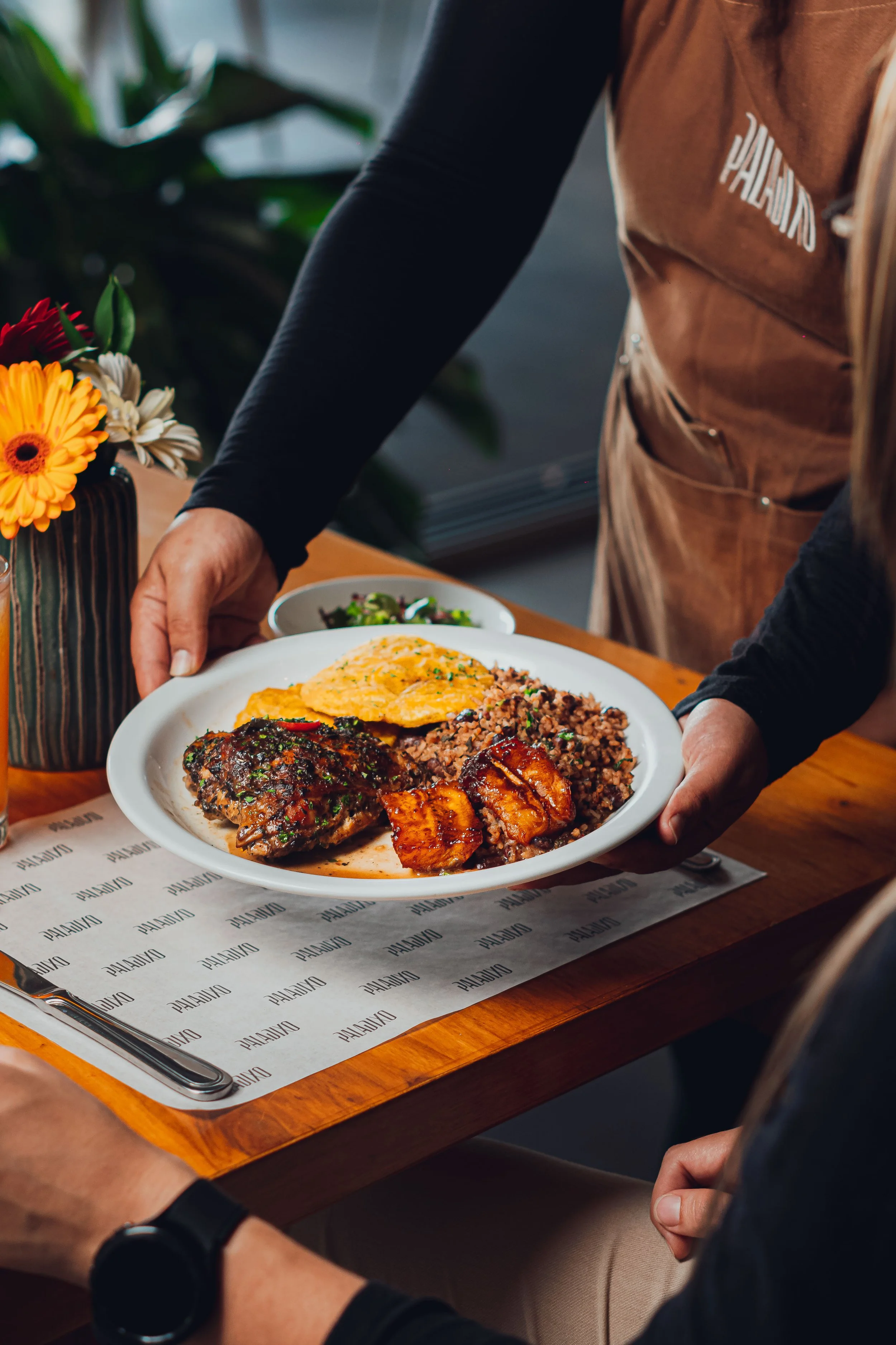 A server is presenting a plate of Mexican food to a customer at a restaurant. The plate contains rice, grilled chicken, cochinita pibil, and a fried tortilla. There is a small bowl of salad in the background.