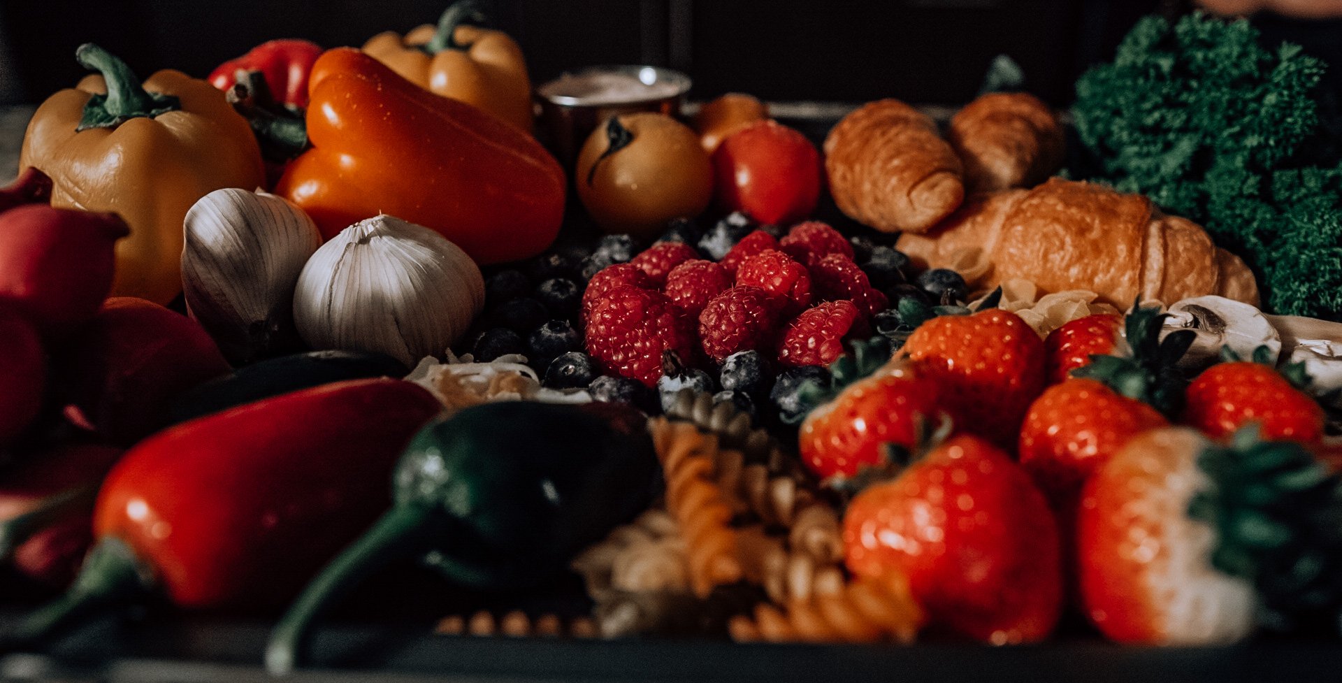 Variedad de frutas y verduras frescas, incluyendo pimientos de colores, ajo, frambuesas, fresas, arándanos, tomates, y vegetales verdes como kale.
