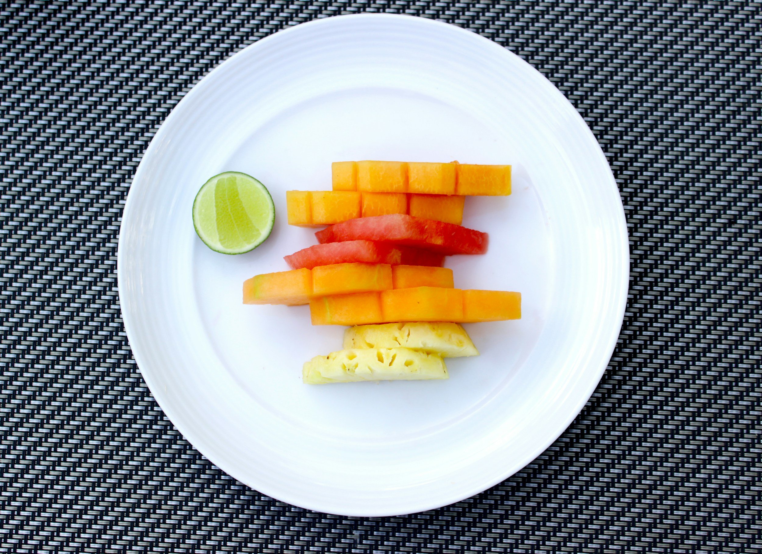 A white plate with sliced watermelon, cantaloupe, pineapple, and lime on a black textured surface.