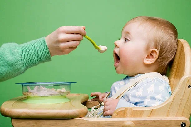 A young child sitting in a wooden high chair with a bib, opening mouth to eat food being fed by a spoon held by an adult's hand, with a green background.