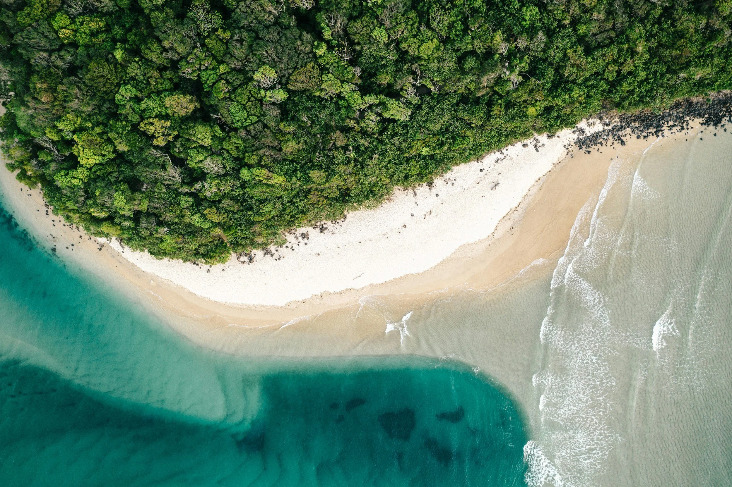 An aerial view of a tropical beach with white sand, surrounded by dense green forest and turquoise ocean water.