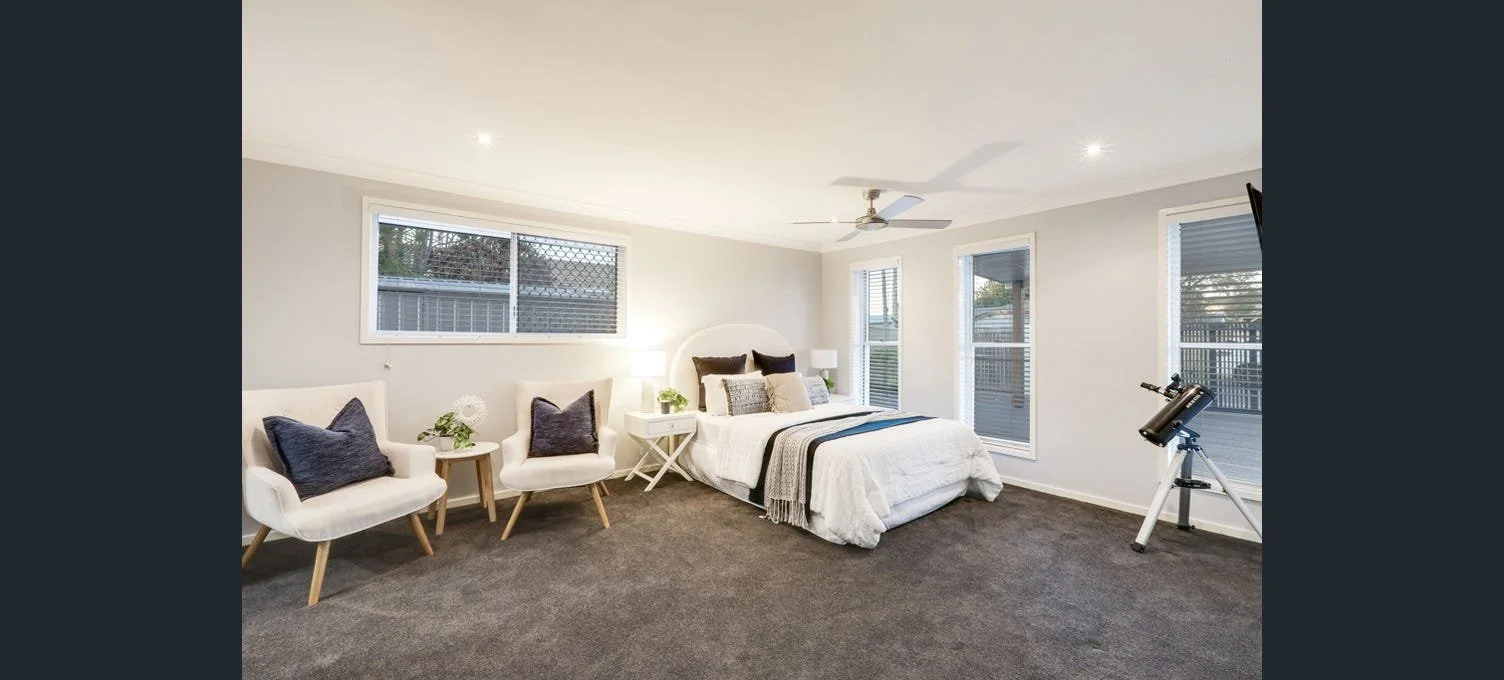 A modern bedroom with a white bed, gray and white bedding, two white nightstands with lamps, two white armchairs with dark pillows, a telescope near the window, and large windows with blinds, carpeted floor, and a ceiling fan.