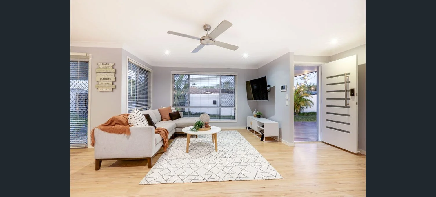 Bright living room with a sectional sofa, white coffee table, TV mounted on the wall, and a door opening to an outdoor area with palm trees.