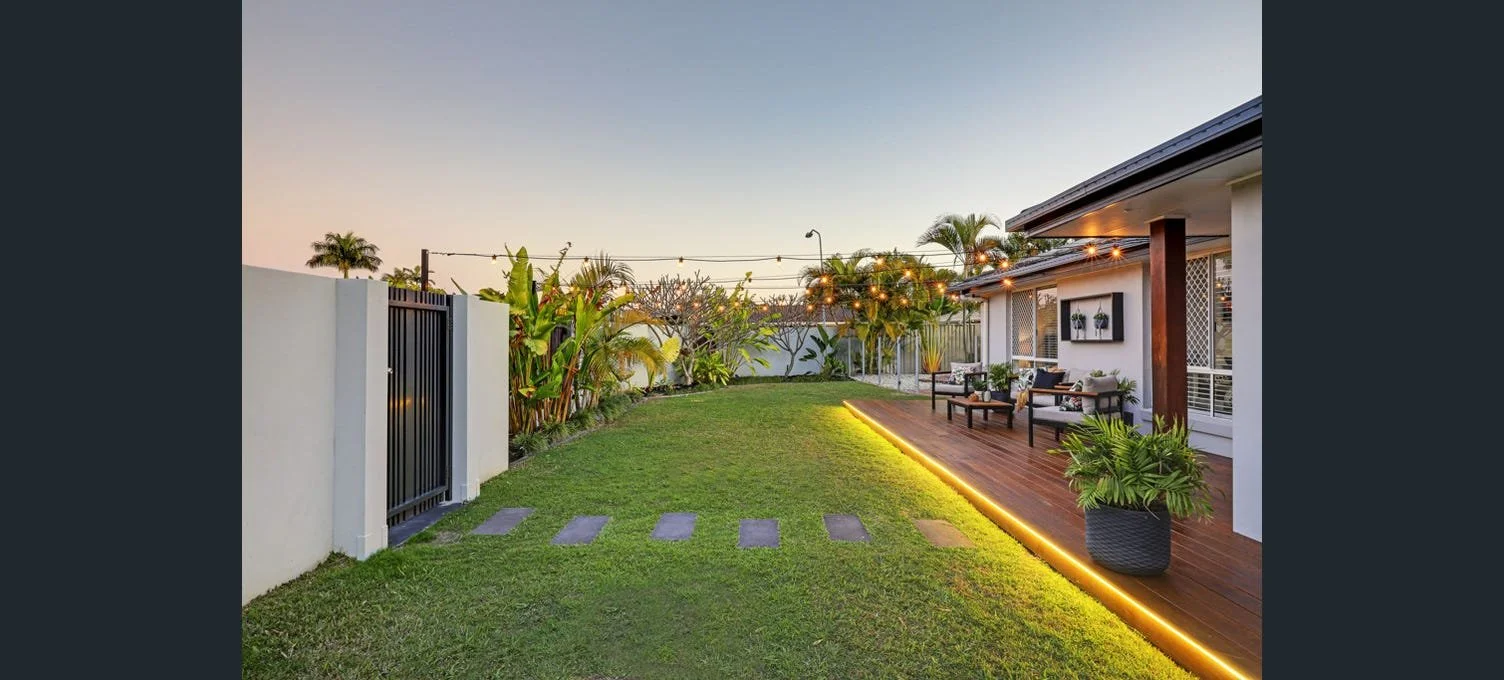 Backyard with a lawn, stepping stones, a wooden deck with outdoor seating, potted plants, string lights, and tropical trees under a sunset sky.