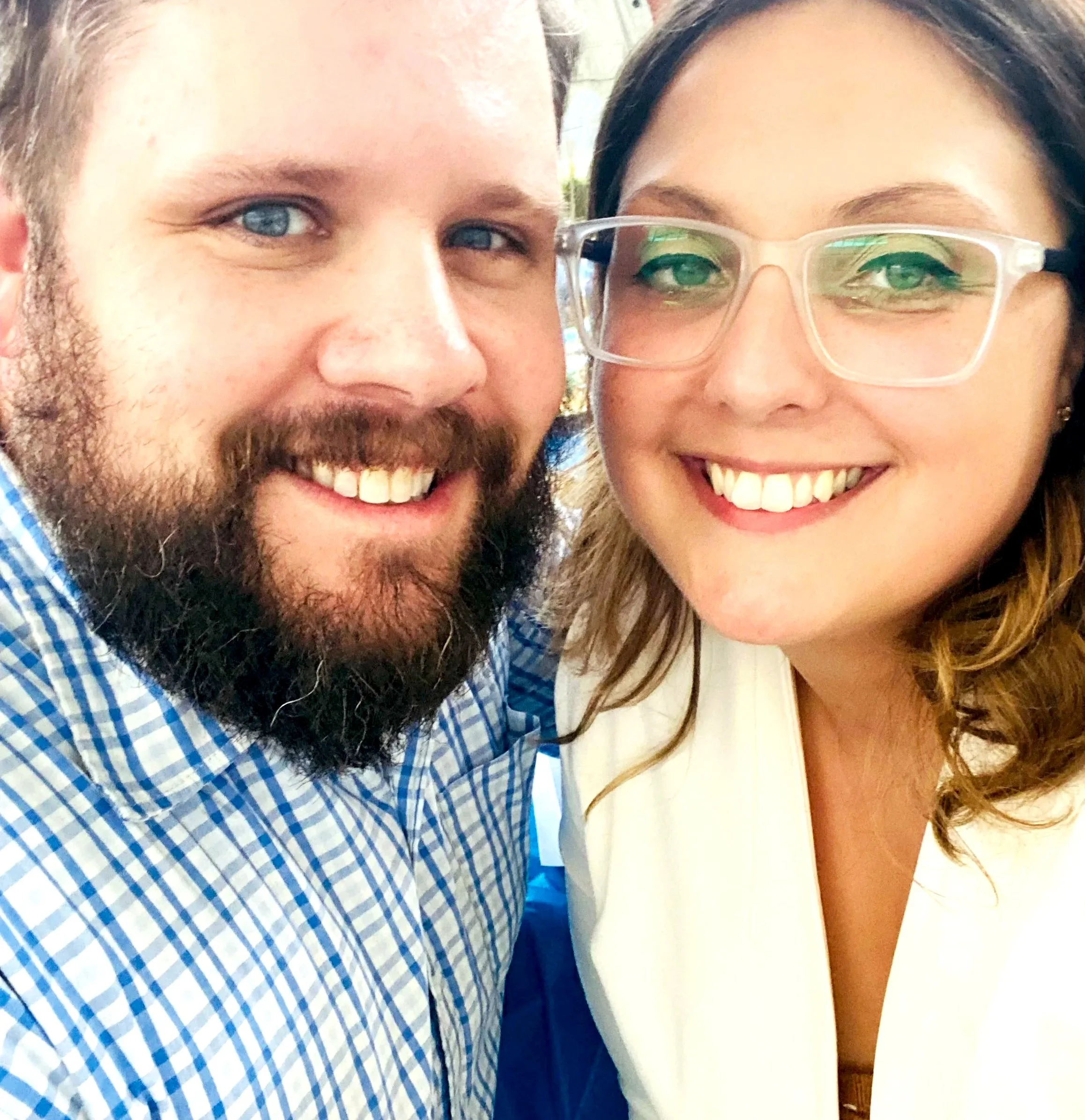 A smiling man with a beard and blue eyes taking a selfie with a smiling woman wearing glasses and a white blazer.
