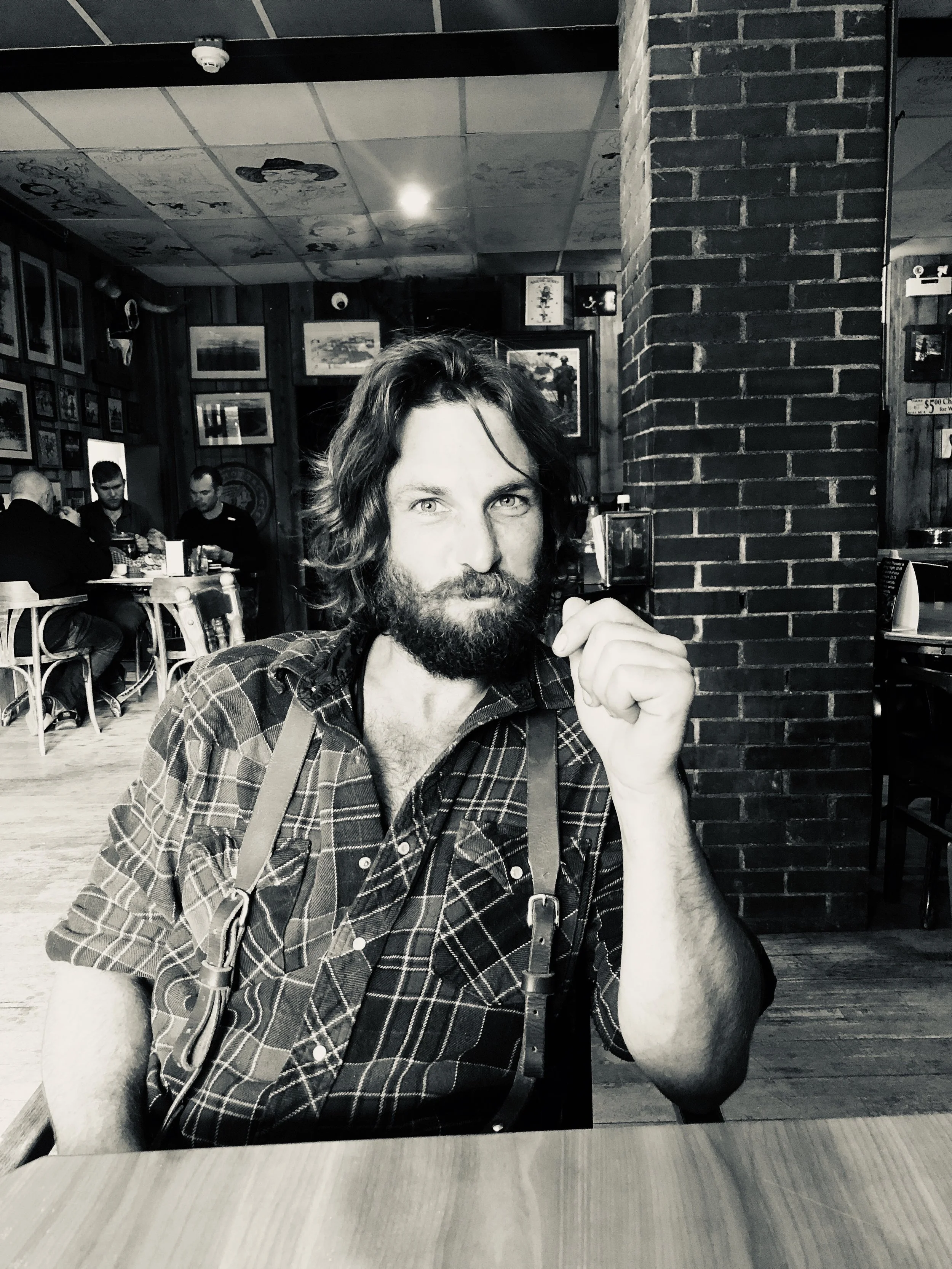 Business owner, carpenter, Black and white photo of a man with a beard and long hair sitting at a table in a restaurant, wearing a plaid shirt and suspenders. He is looking directly at the camera with a thoughtful expression.
