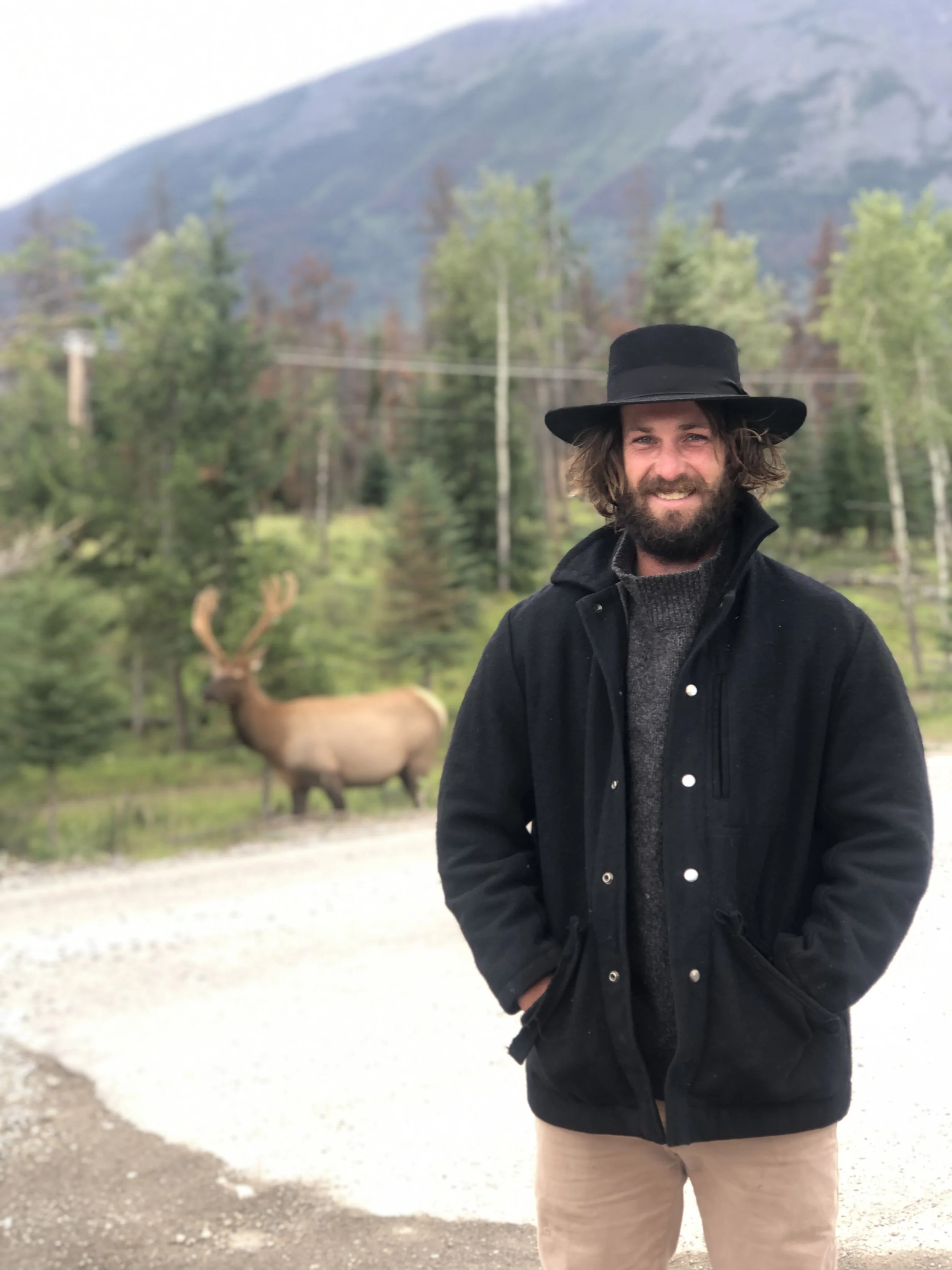 Business owner, carpenter, a man with a beard and long hair wearing a black hat, black jacket, and beige pants standing outdoors on a gravel road, with a moose in the background among trees and mountains.