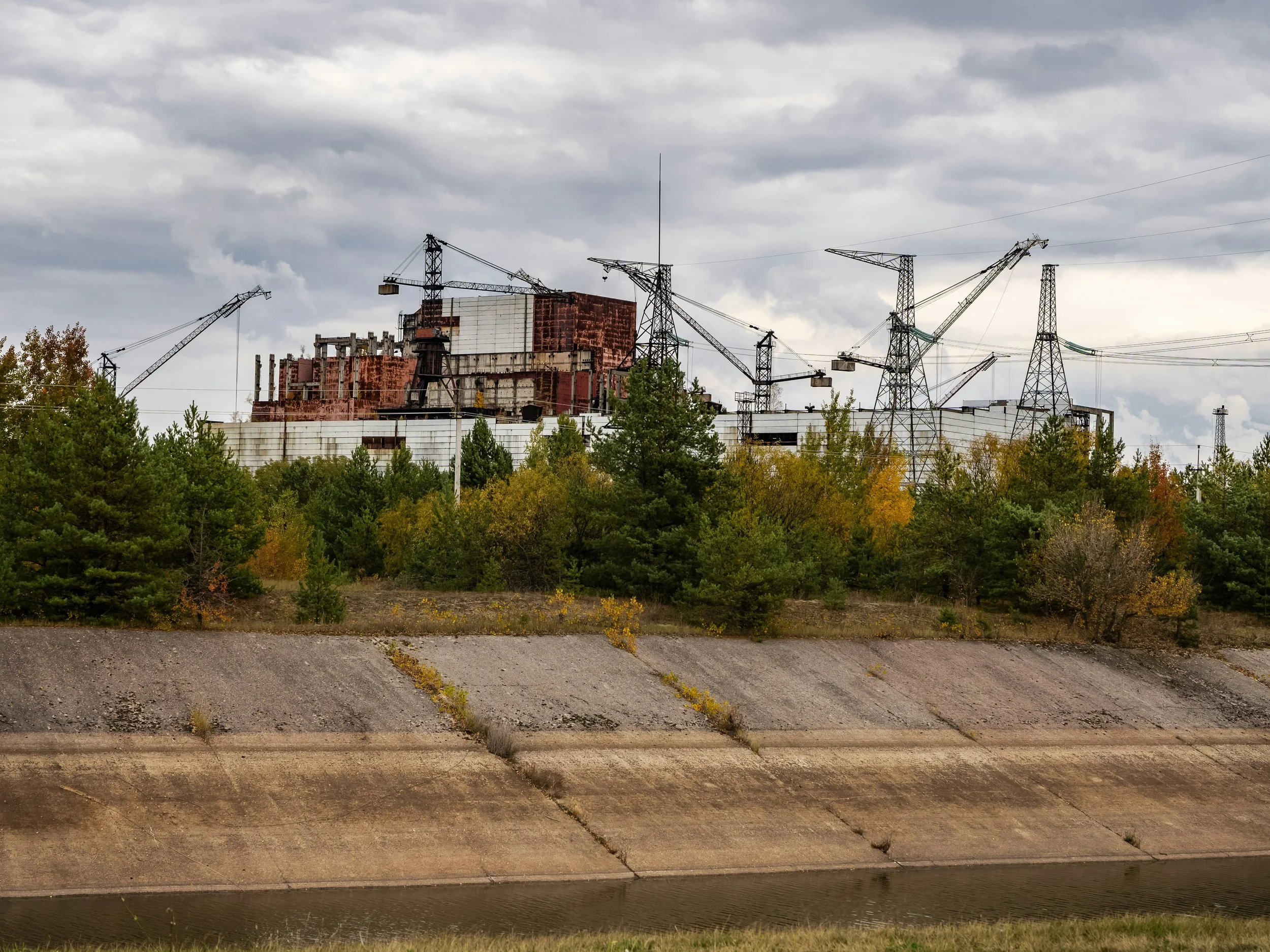 Industrial building under construction with multiple cranes, surrounded by trees and a concrete embankment, under a cloudy sky.