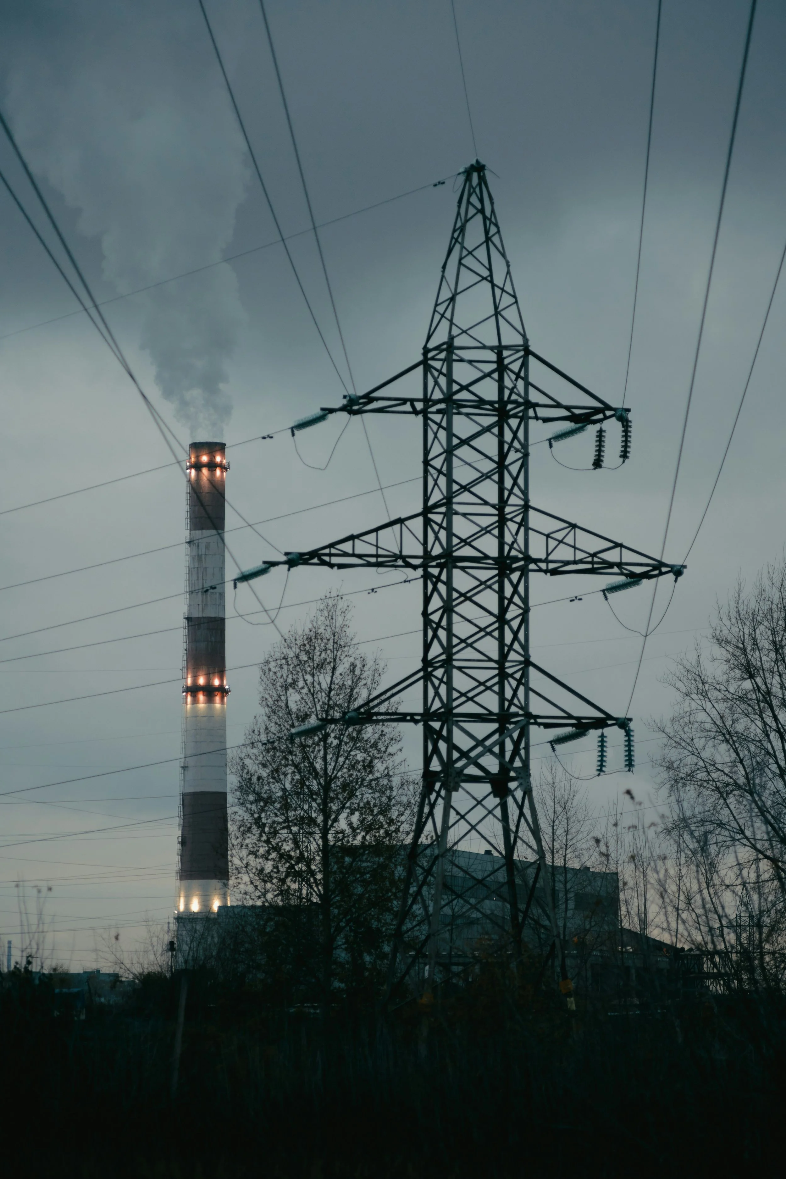 Electrical transmission tower with power lines, factory smokestack emitting smoke in the background, and trees in a gloomy sky.