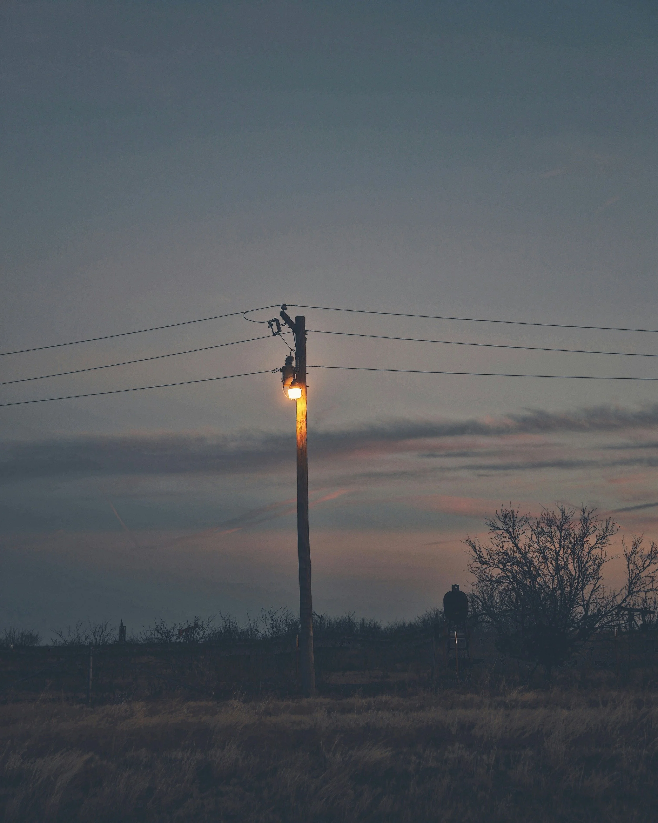 A utility pole with a streetlamp on at dusk, with a dark cloudy sky and barren trees in the background.