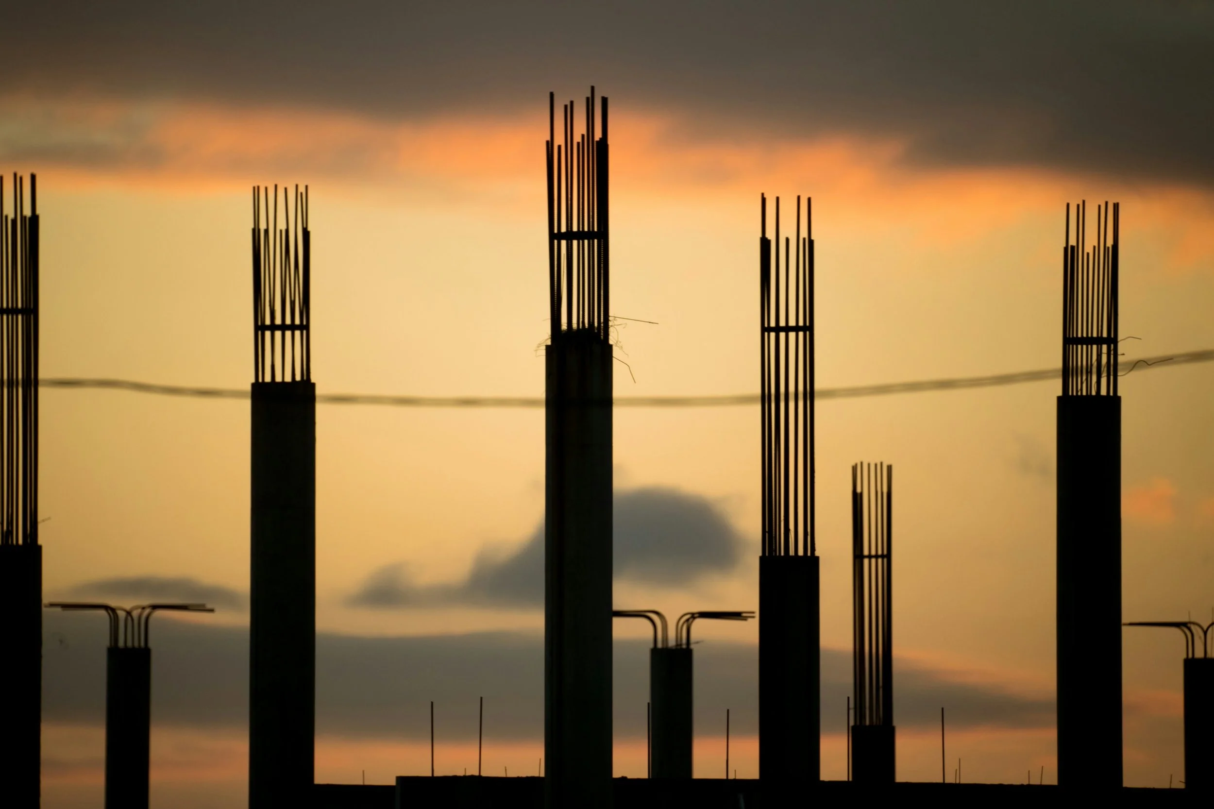Silhouette of unfinished building columns with steel rebars against a sunset sky with clouds.