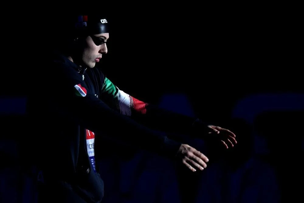 Atleta in abbigliamento nero con patch dell'Italia, indossa occhiali e cuffia, all'aperto al buio, si prepara probabilmente per una gara o allenamento.