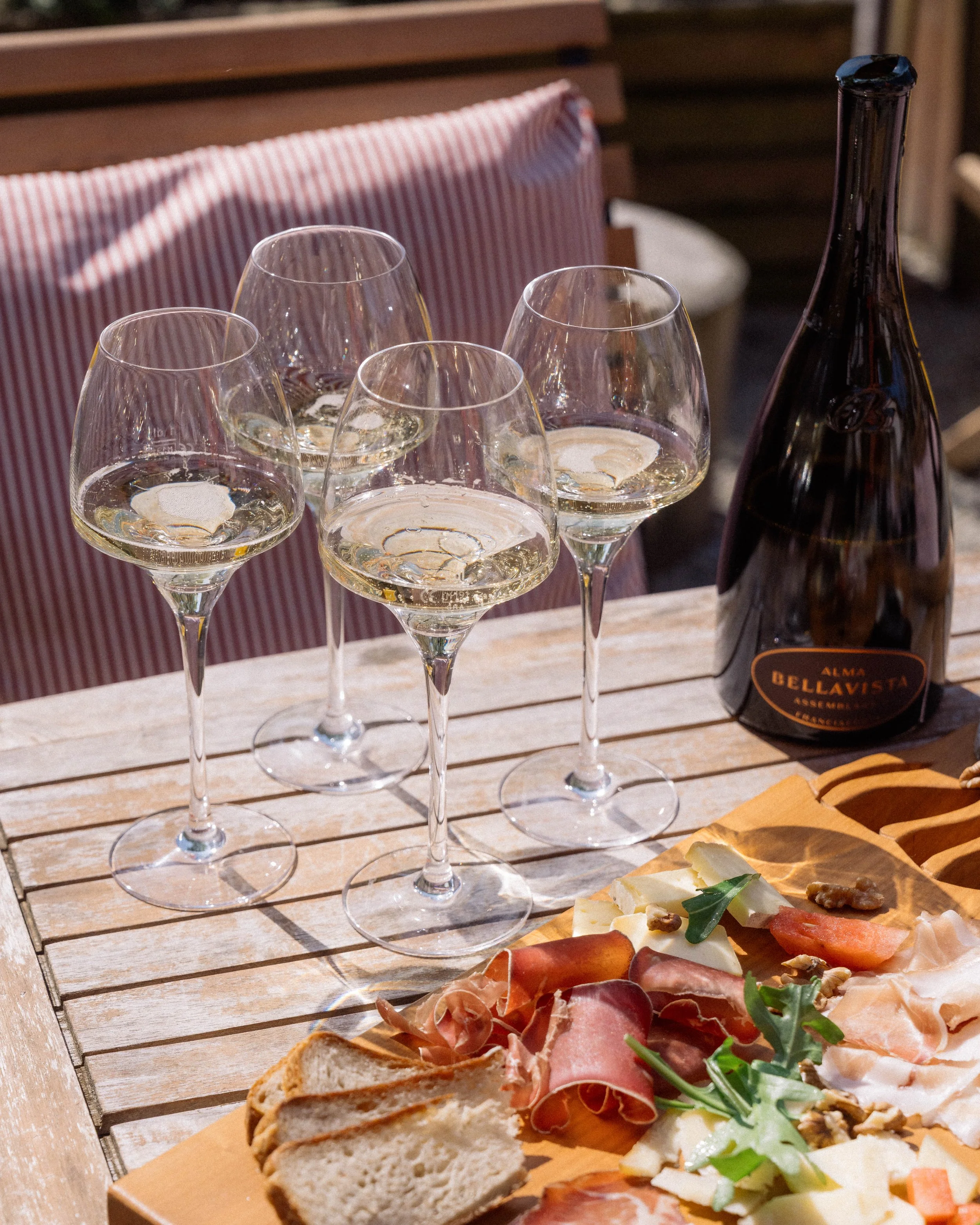 Vignette d'une table avec quatre flûtes de champagne, une bouteille de champagne Alma Bellavista, un plateau de fromages, charcuterie, noix et pain, dans un cadre en plein air