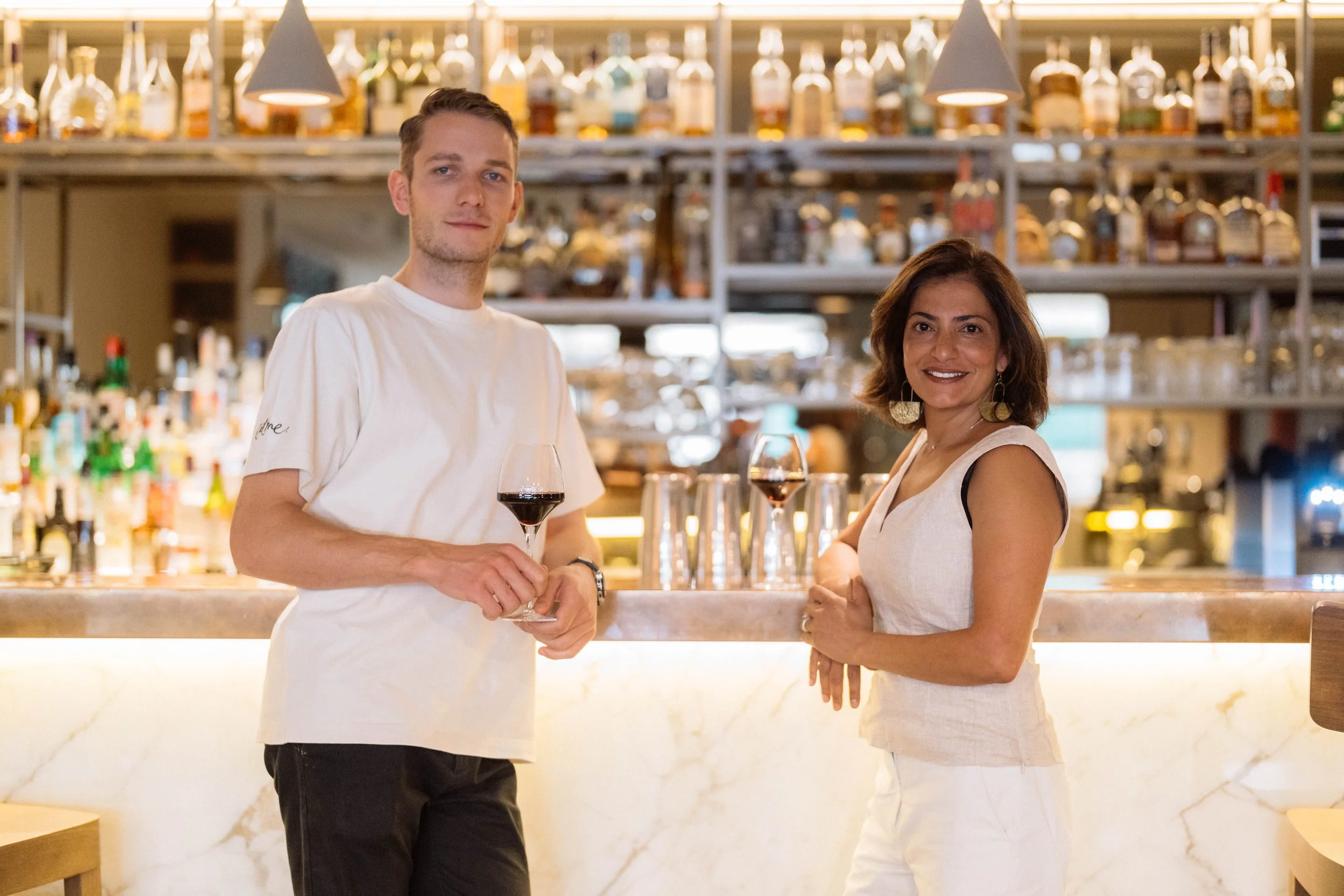Un homme et une femme souriants, debout au bar, tenant chacun un verre de vin rouge, dans un bar avec étagères remplies de bouteilles d'alcool en arrière-plan.