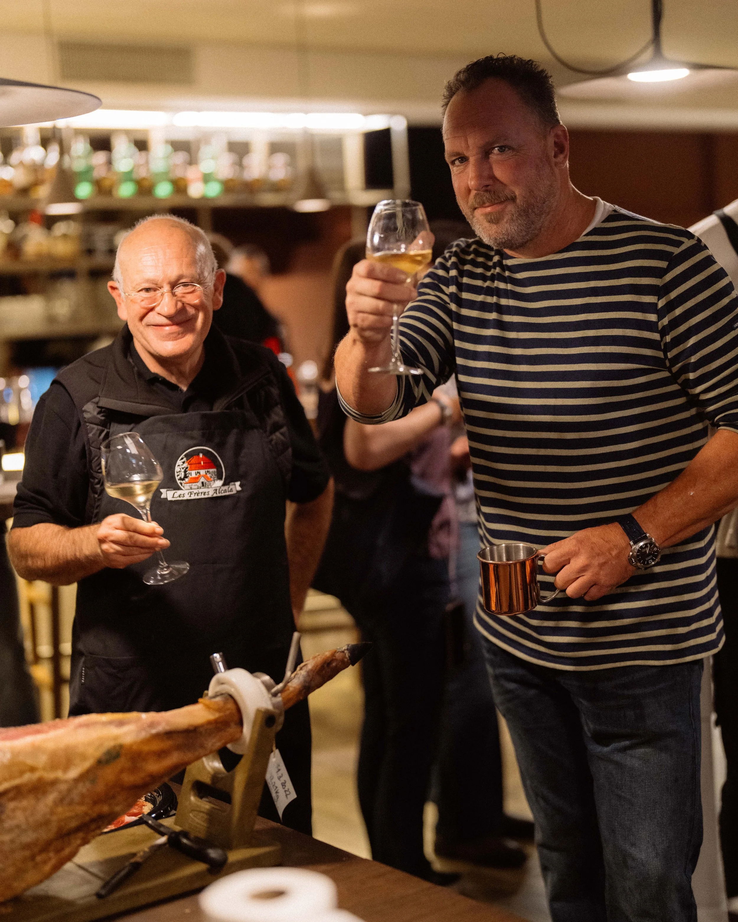 Deux hommes dans un restaurant, l'un portant un tablier, tenant un verre de vin blanc, l'autre tenant un verre de vin, portant un mug. Il y a un jambon suspendu sur une machine à couper devant eux.