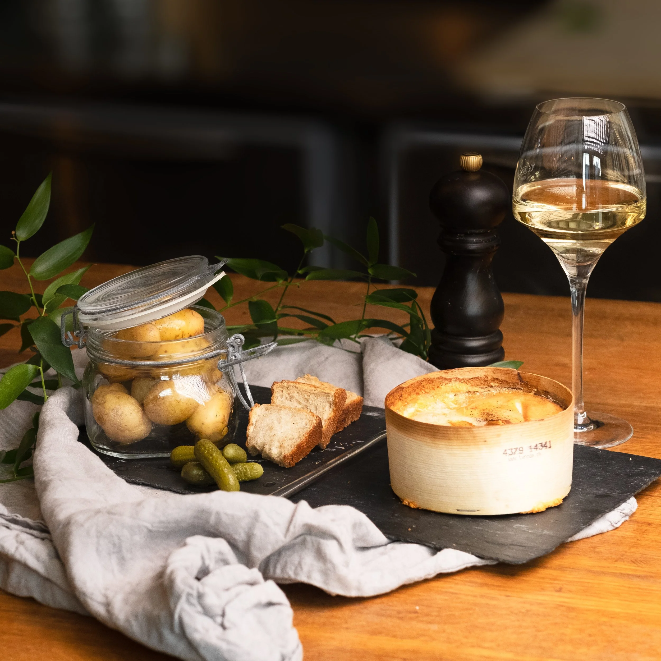 Plat de fromage avec des gressins, des pommes de terre en bocal, des cornichons, un verre de vin blanc, un ramequin de fromage fondu, et un moulin à poivre sur une table en bois.
