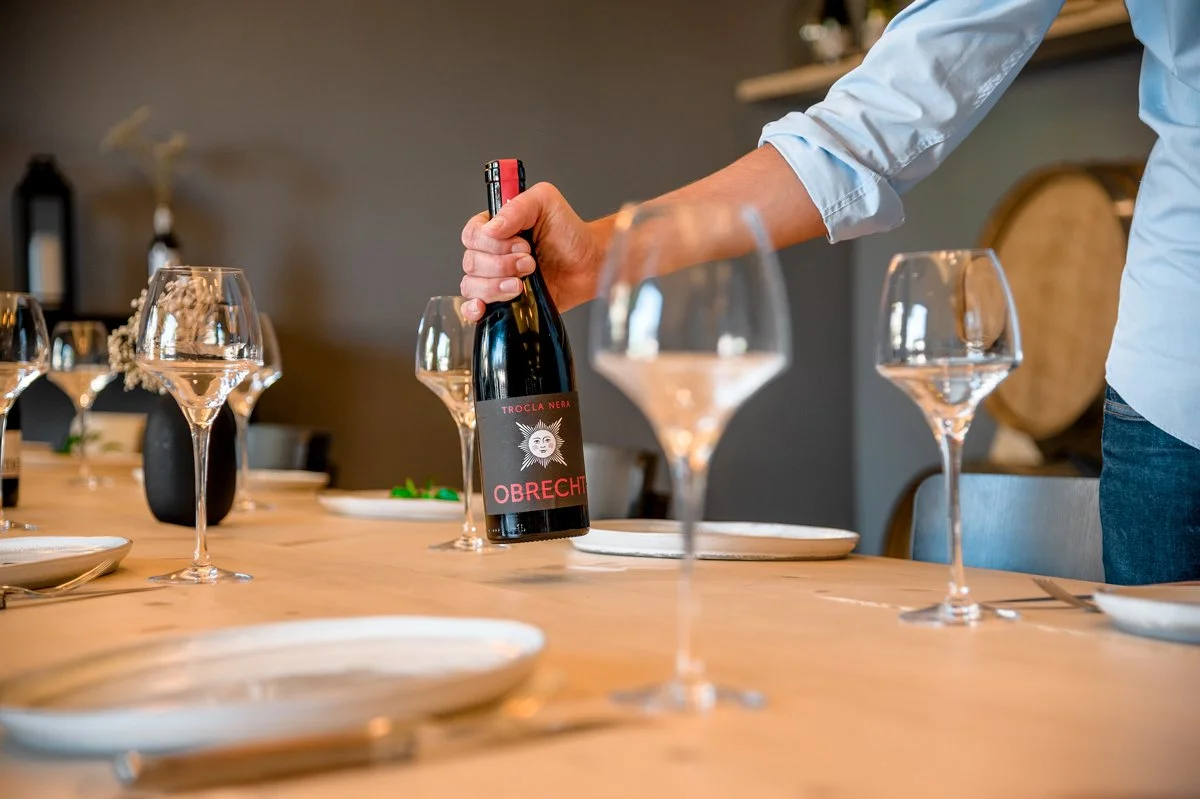 Une personne dévisse une bouteille de vin rouge, qui porte l'étiquette 'Obrecht'. Il y a plusieurs verres à vin et assiettes vides sur une table en bois, prêts pour un repas.