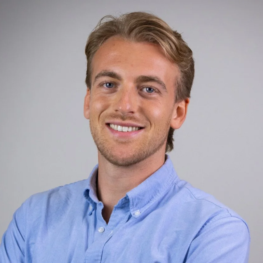 Portretfoto van een lachende jonge man met blond haar, blauwe ogen en een lichtblauw overhemd, staand voor een neutrale grijze achtergrond.
