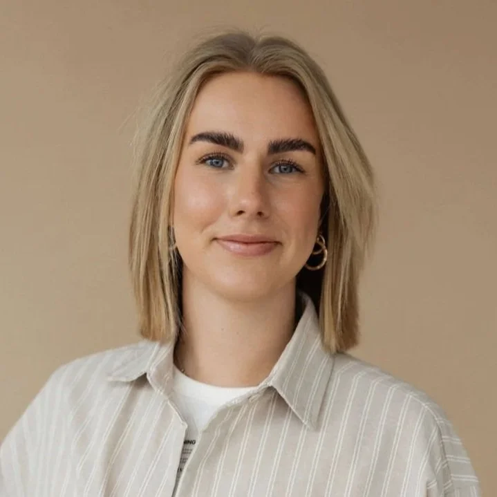 Portret van een jonge vrouw met blonde haar, blauwe ogen, licht getinte huid, gekleed in een wit shirt met streeppatroon, tegen een beige achtergrond.