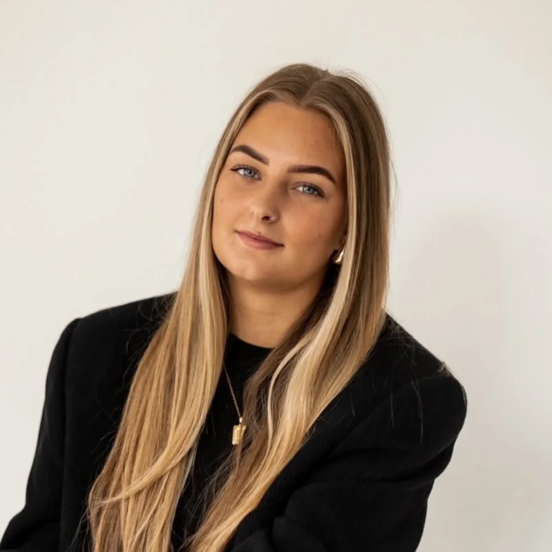 Portret van een jonge vrouw met lang blond haar, blauwe ogen en een neuspiering, gekleed in een zwarte jas, staand tegen een witte muur.