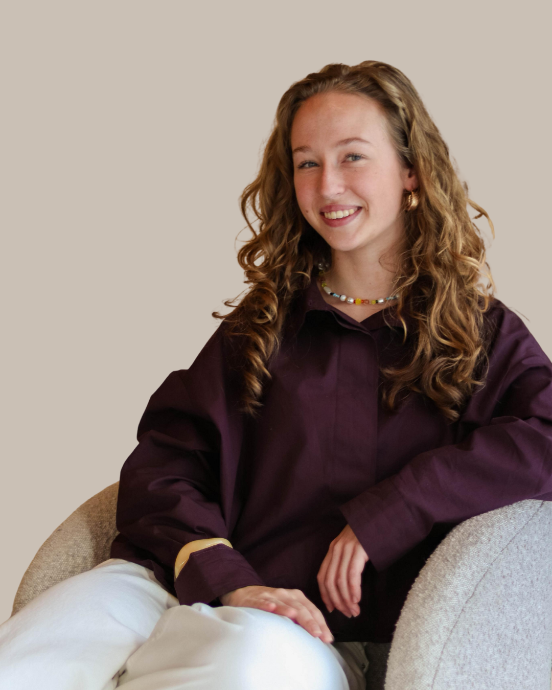 een jonge vrouw met krullend haar, lachend, zittend op een beige stoel, gekleed in een paarse blouse en witte broek, met een kleurrijke ketting en gouden oorbellen.