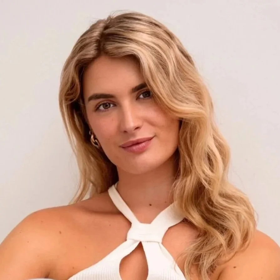 Portret van een vrouw met lang, golvend blond haar, lichte huidskleur en een witte top, glimlachend tegen een neutrale achtergrond.