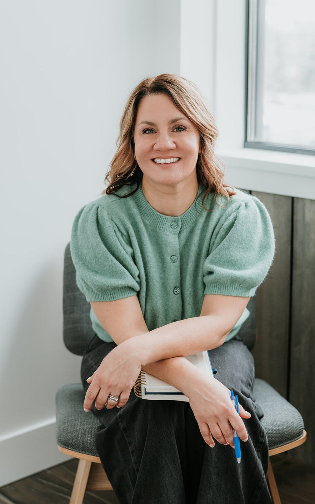 A woman with shoulder-length light brown hair wearing a green short-sleeve button-up sweater, sitting on a gray chair near a window, holding a notebook and blue pen, smiling at the camera.