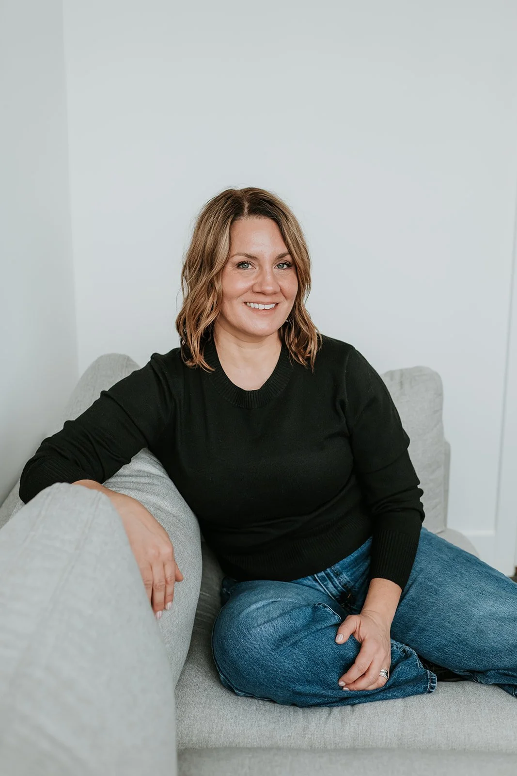 A woman with shoulder-length wavy brown hair, wearing a black sweater and blue jeans, sitting on a light gray sofa in a minimalistic room with white walls and smiling at the camera.
