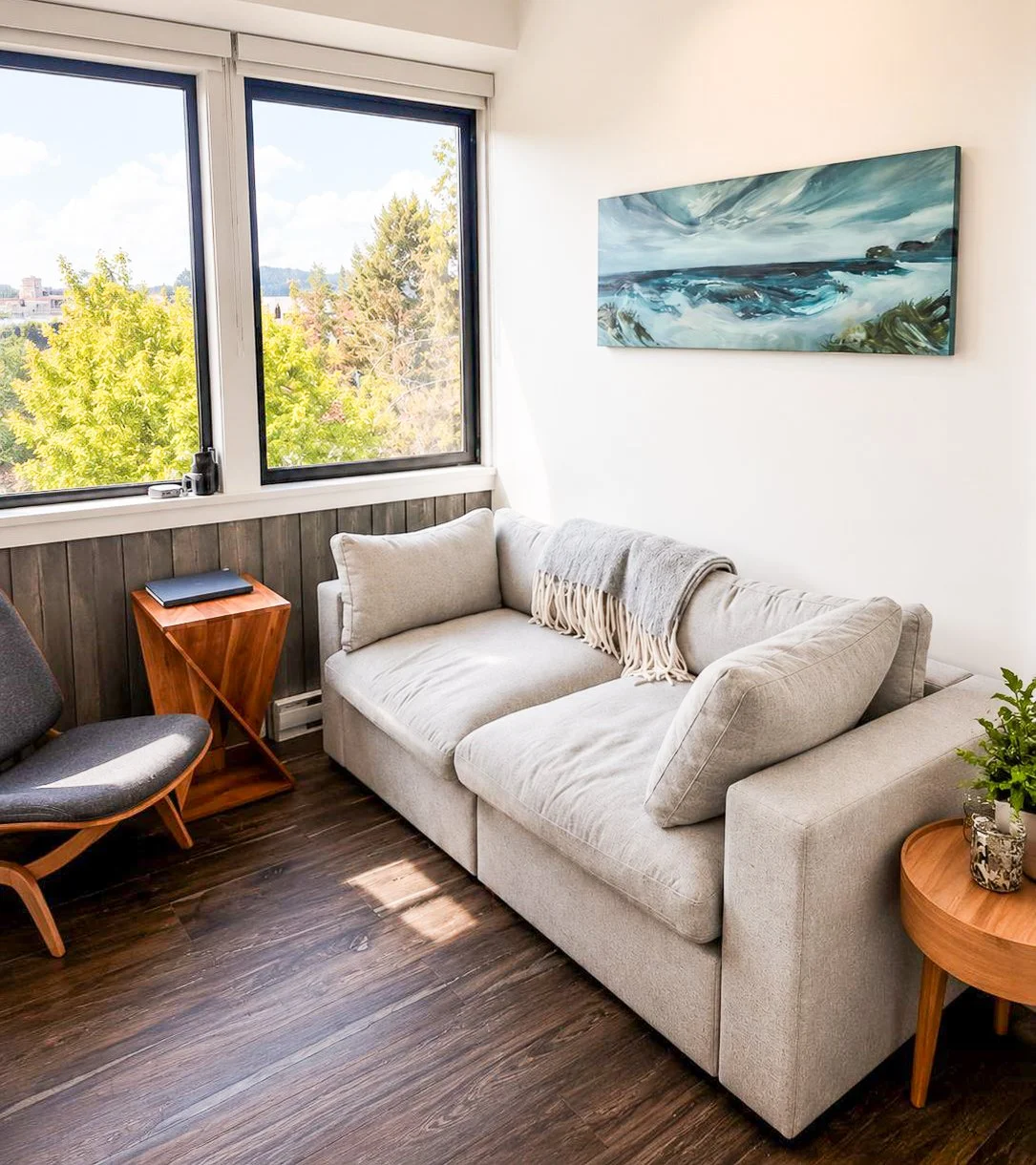 Living room with large window showing green trees outside, beige sofa with throw blanket, wooden side table, wall art depicting a seascape, gray chair, and a small potted plant.