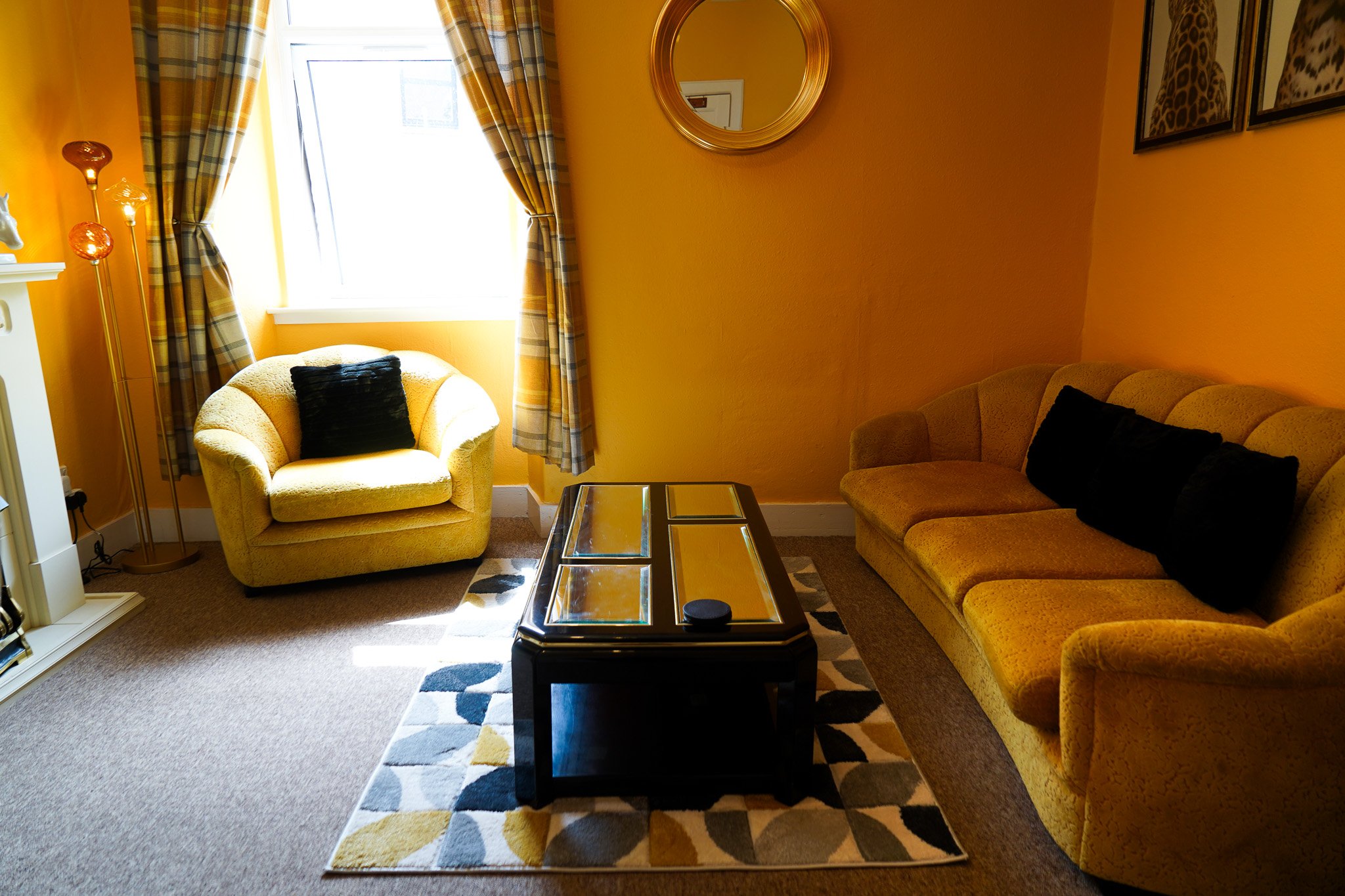 Living room with yellow walls, a window with plaid curtains, a yellow armchair with a black pillow, a yellow sofa with black pillows, a black coffee table with glass panels, a patterned rug, and framed pictures on the wall.