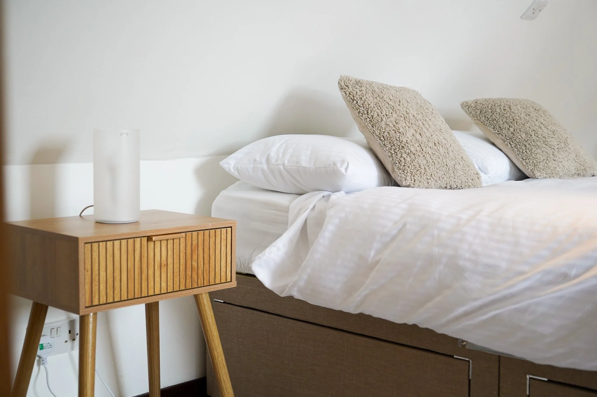 Close-up of a bed with white bedding and three beige textured pillows, alongside a wooden bedside table with a white diffuser, in a minimalistic bedroom.