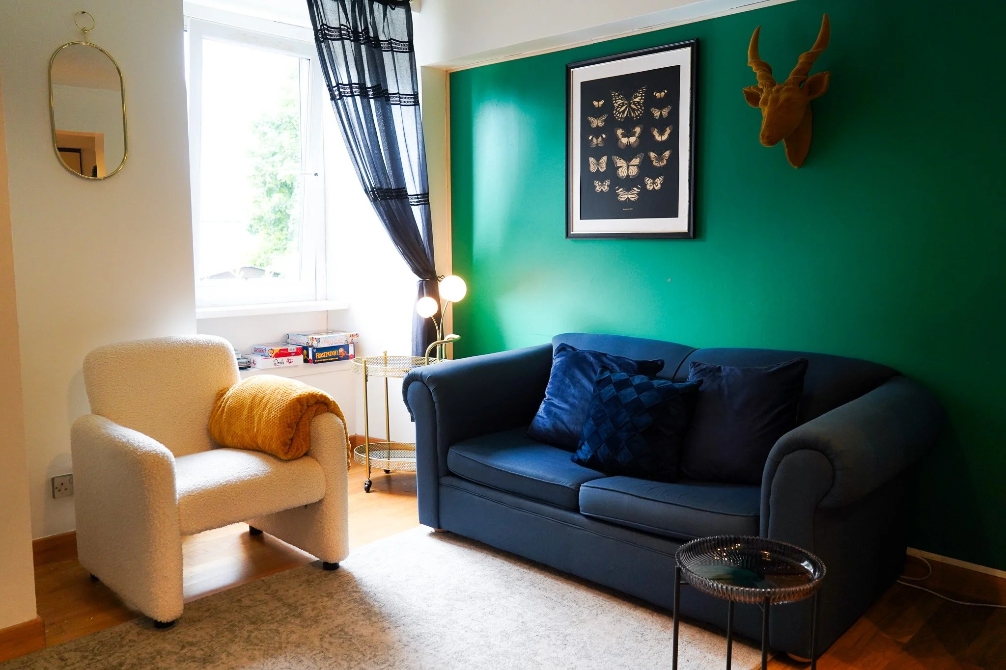 Living room with a green accent wall, dark blue sofa with cushions, white armchair with a yellow blanket, window with dark curtains, framed butterfly artwork, deer head wall decor, side table, floor lamp, and a window seat with board games.