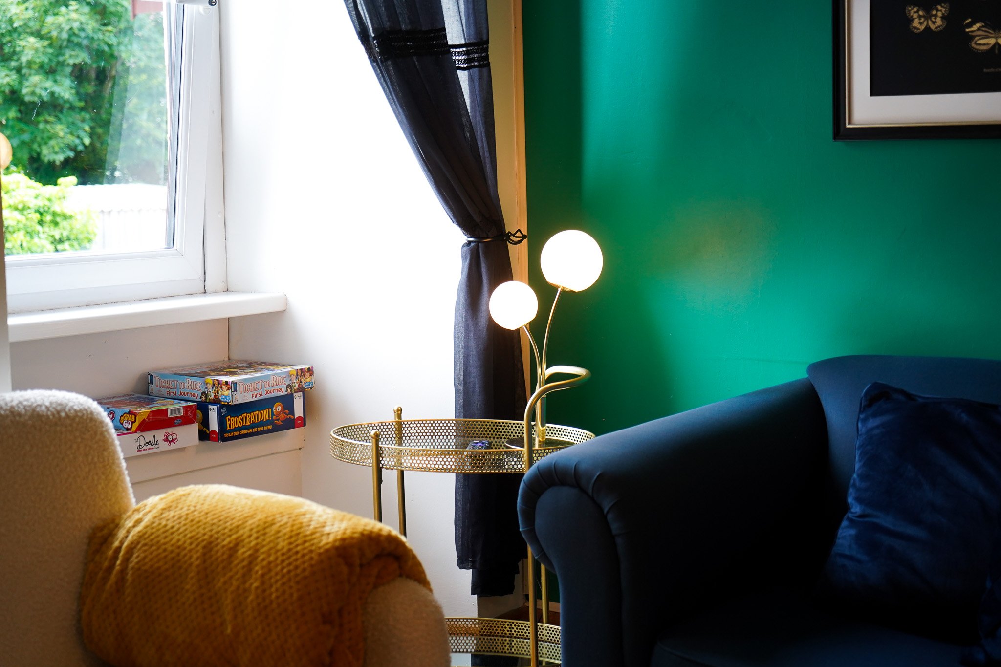 Living room corner with a dark blue couch with a pillow, a yellow armchair with a knitted yellow throw, a gold side table, and a modern gold and white floor lamp. There is a green accent wall, artwork on the wall, a window with dark curtains, and board games on the windowsill.