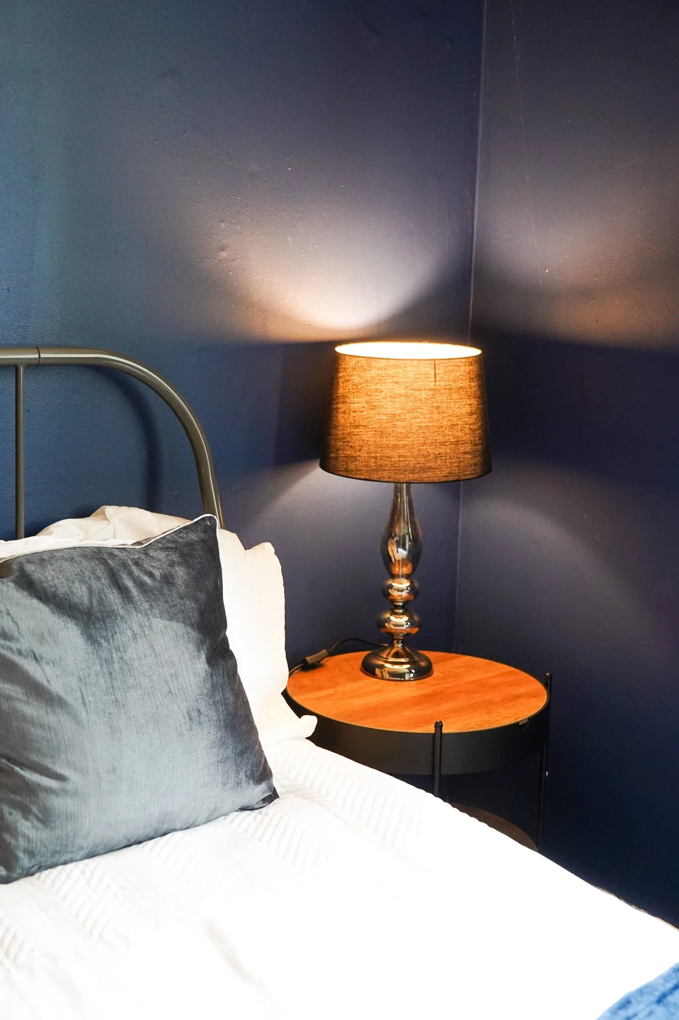 A bedroom corner with a bed, decorative pillows, a round wooden bedside table, and a table lamp with a black shade on a dark blue wall.