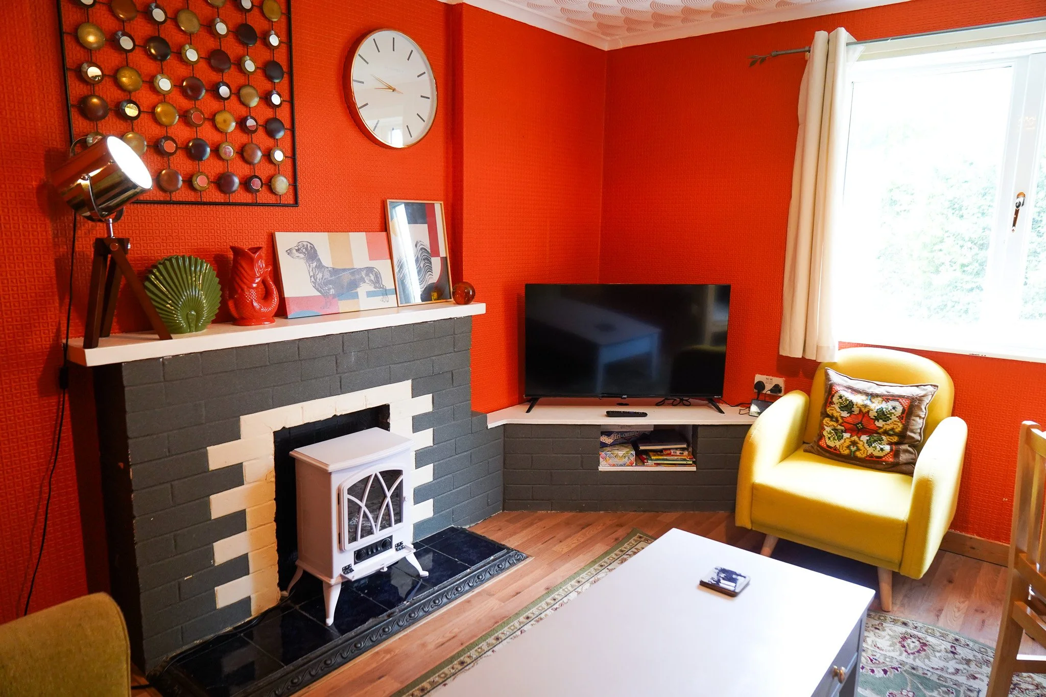 A living room with orange textured walls, a black and white fireplace, a yellow armchair with a floral pillow, a flat-screen TV on a corner shelf, a window with white curtains, a large wall clock, and decorative items on the mantel.